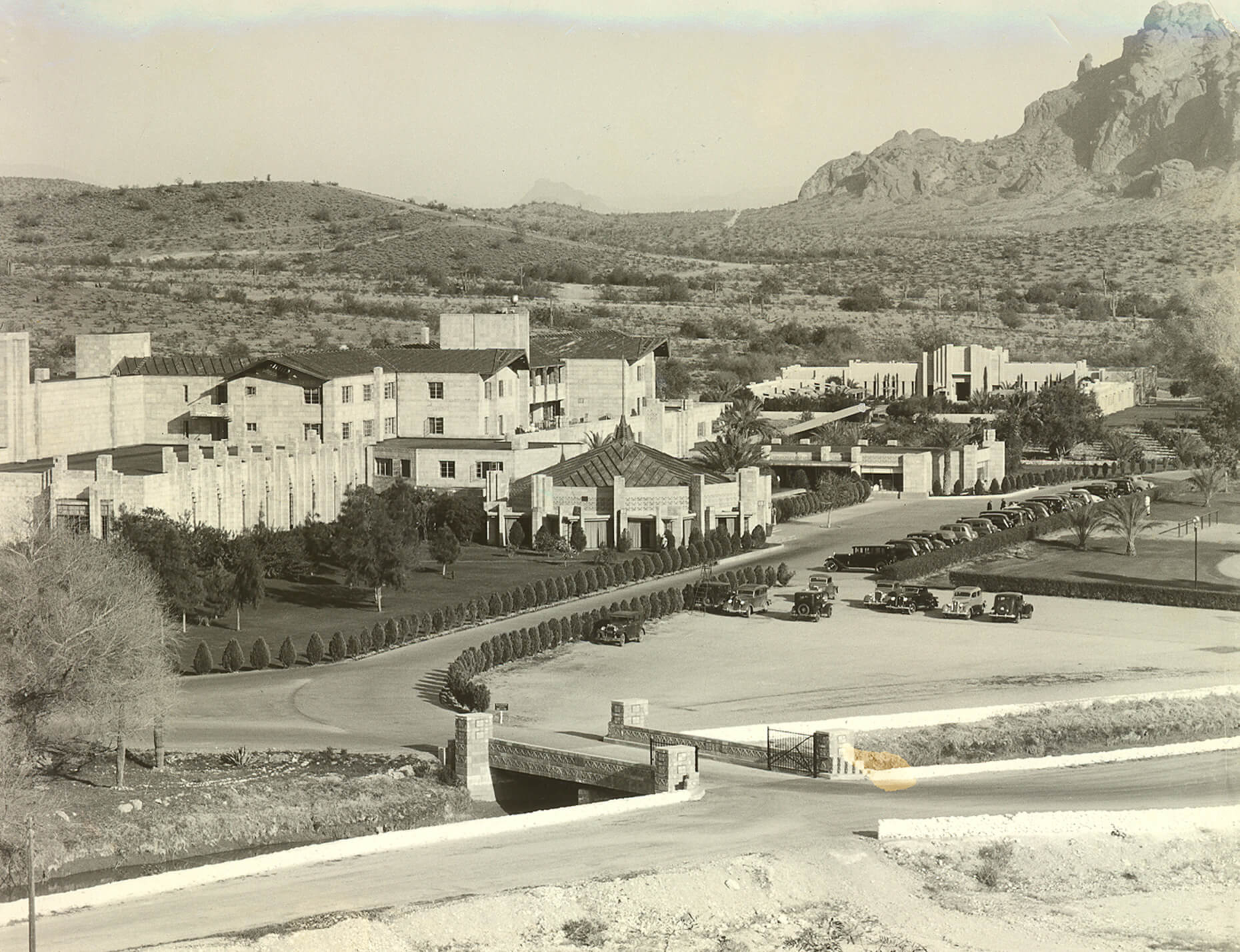A sepia-toned image depicting a large, sprawling building complex with a desert landscape and mountain in the background. The entrance features a row of neatly trimmed bushes, showcasing meticulous preservation, and numerous vintage cars are parked along the driveway.