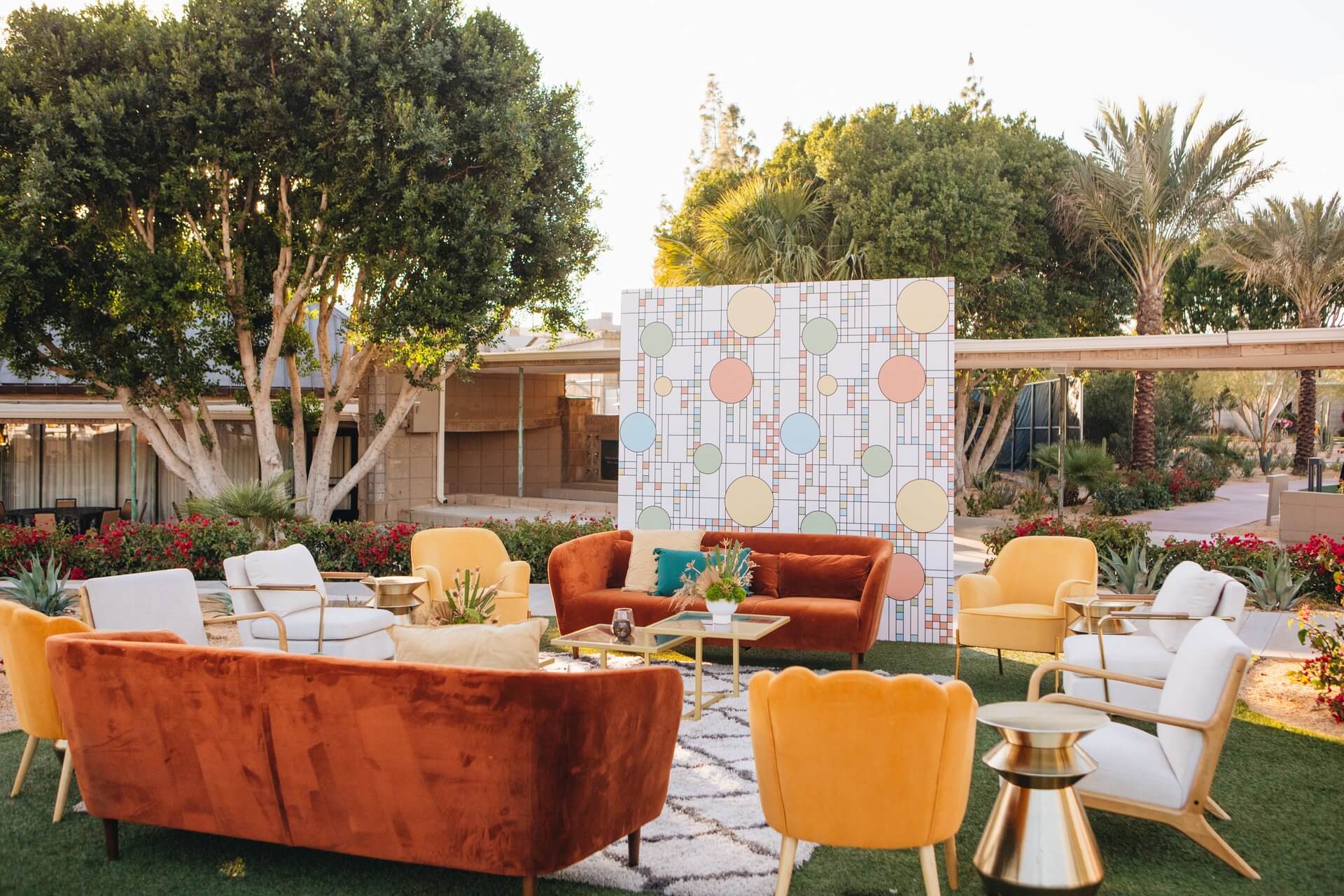 An outdoor seating area features a mix of colorful furniture, including orange, white, and yellow chairs and sofas. The setup includes a geometric backdrop with circles and lines, surrounded by lush greenery and palm trees, creating a modern and inviting atmosphere perfect for meetings & events.