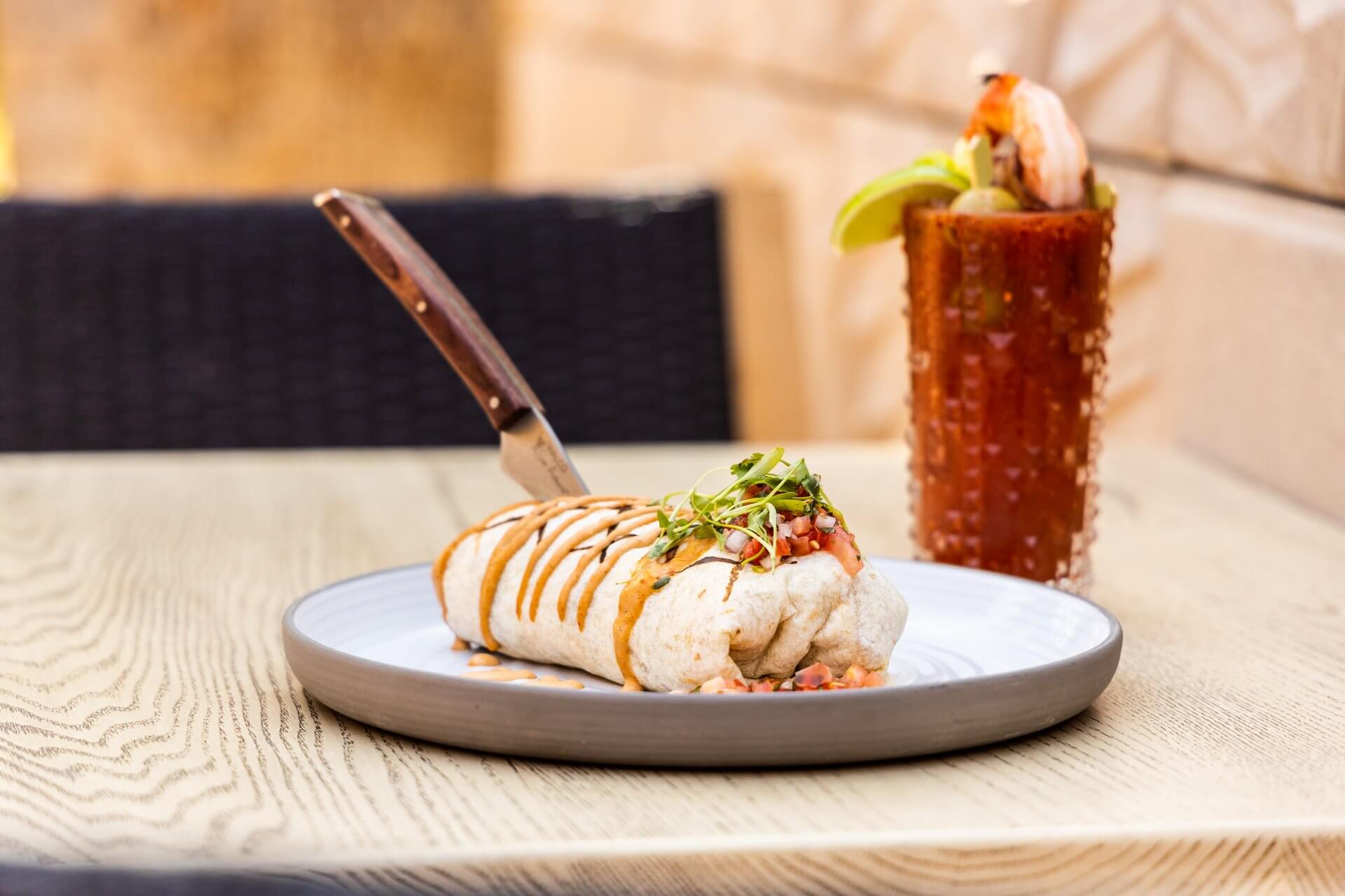 A burrito is placed on a white plate at McArthur's, garnished with sauce and microgreens, with a knife sticking out from the top. In the background, there is a tall cocktail glass filled with a shrimp-topped beverage and a lime wedge on the rim, resting on a wooden table.