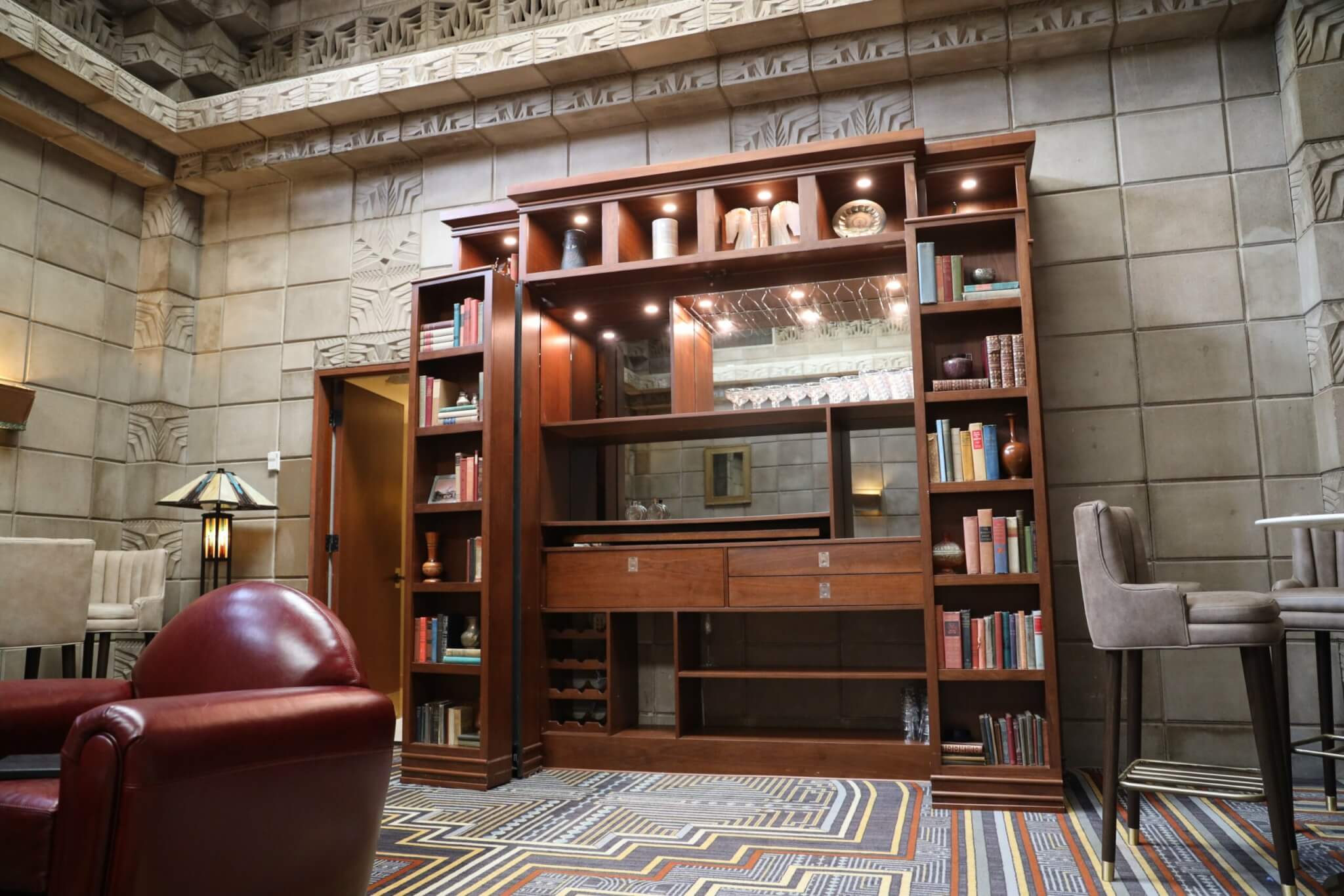 A cozy, modern library with wooden bookshelves filled with books and decorative items. A leather armchair is in the foreground, and a high table with stools is on the right. The room features intricate wall designs, a patterned carpet, and an air of a mystery room waiting to be explored.