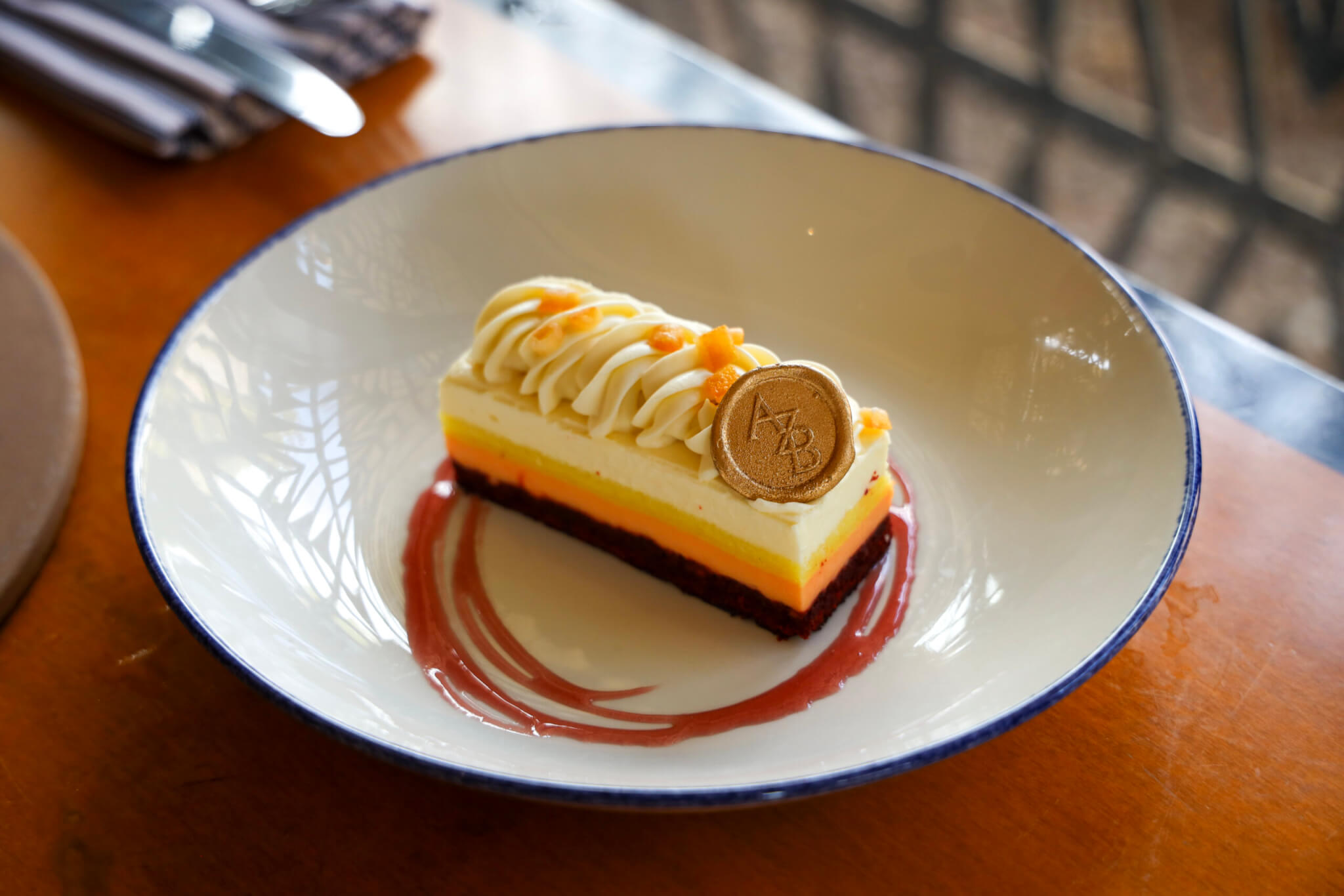 A plated rectangular dessert with layers of yellow, orange, and white, topped with piped cream, orange zest, and a round chocolate disc, served in a white bowl with a red sauce swirl.