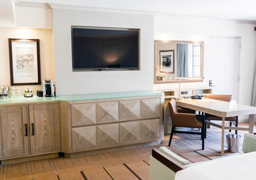 A well-lit hotel room, part of the Citrus Club Balcony suite, features a large flat-screen TV mounted on a wall above a wooden cabinet. The room includes a desk with an orange chair, a coffee maker with cups, a mirror above the desk, and framed artwork on the wall. The window is partially covered by curtains.