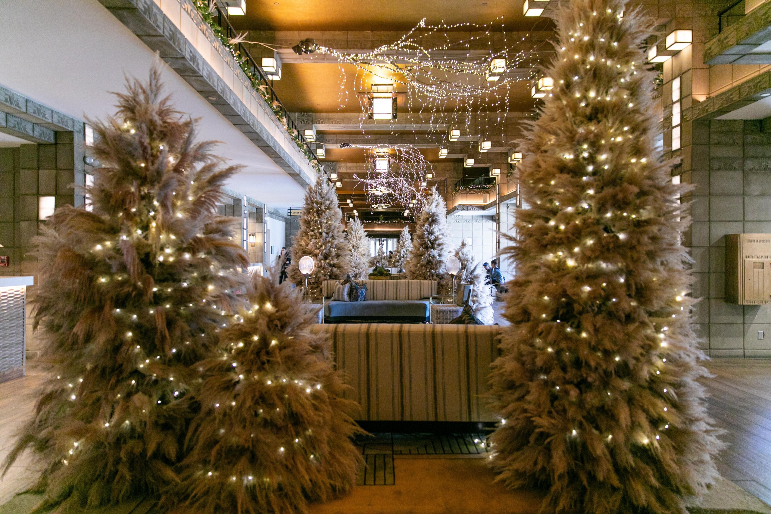 A spacious, elegant hall decorated with large pampas grass Christmas trees adorned with white string lights, soft seating, and warm ceiling lights, creating a festive and cozy atmosphere—perfect inspiration for The Ultimate Gift Guide.