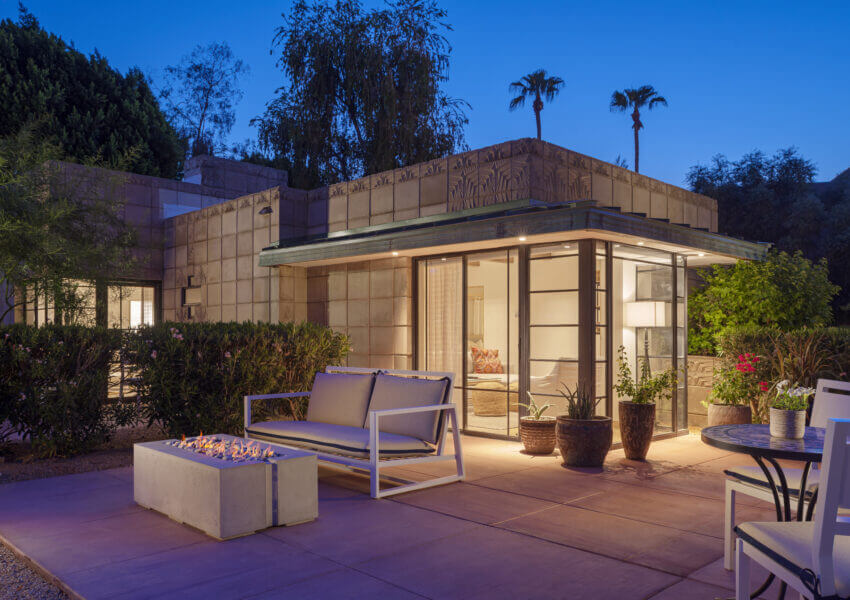 A modern outdoor patio at dusk, featuring a cozy seating area with a white cushioned bench and chairs surrounding a contemporary gas fire pit. The scene resembles a luxurious cottage suite, with the background showcasing a geometric-styled house and lush greenery framing the well-lit interior.