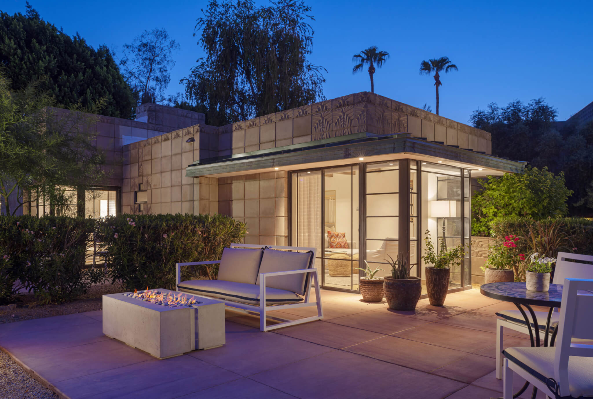A modern outdoor patio at dusk, featuring a cozy seating area with a white cushioned bench and chairs surrounding a contemporary gas fire pit. The scene resembles a luxurious cottage suite, with the background showcasing a geometric-styled house and lush greenery framing the well-lit interior.