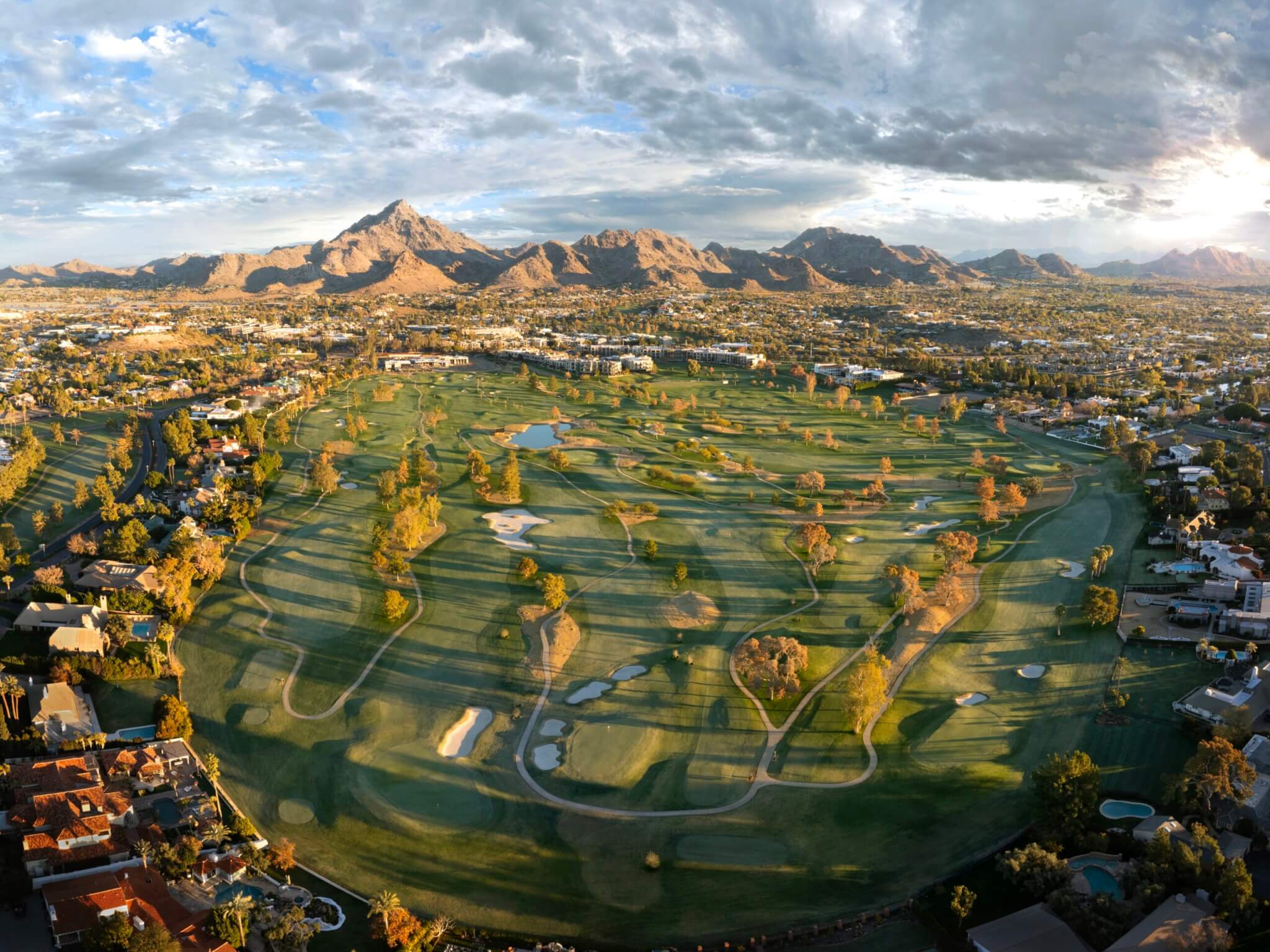 Aerial view of a lush green golf course surrounded by residential areas and a mountainous landscape in the background. The sky is partly cloudy, with sunlight casting shadows across the terrain, highlighting the various golf holes, sand traps, and water hazards—a golfer's paradise.