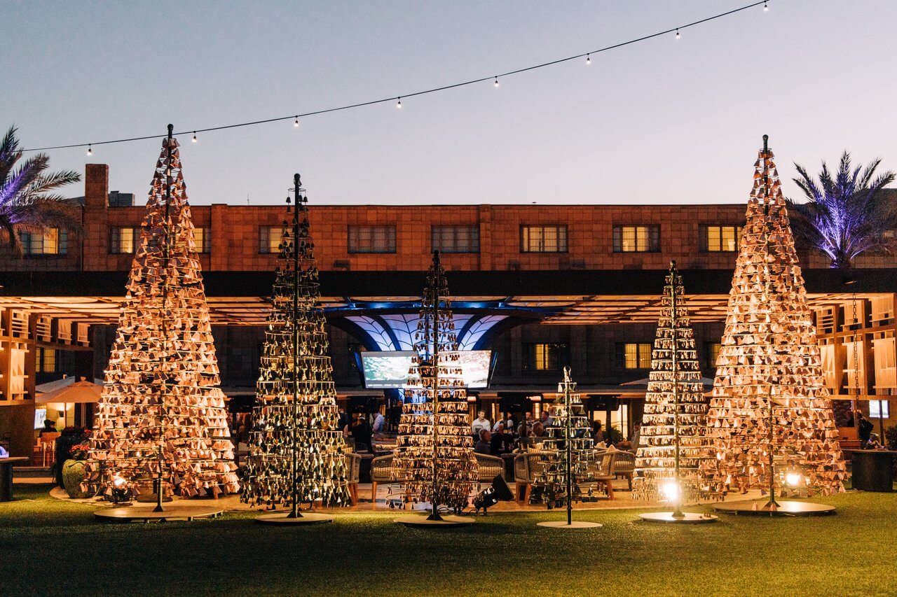 A festive outdoor scene at dusk features several tall, illuminated triangular structures resembling Christmas trees. People gather around, enjoying Hot Rates on cool dates, while a large building with a patio and string lights adds to the enchanting atmosphere in the background.