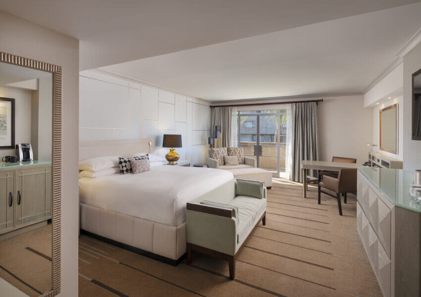 A spacious, well-lit hotel room featuring a king-sized bed with white linens, a cushioned headboard, and a small bench at the foot. The room includes a sitting area with a sofa, a desk, a large window, a wall-mounted TV, and a coffee station. The decor is modern and neutral.