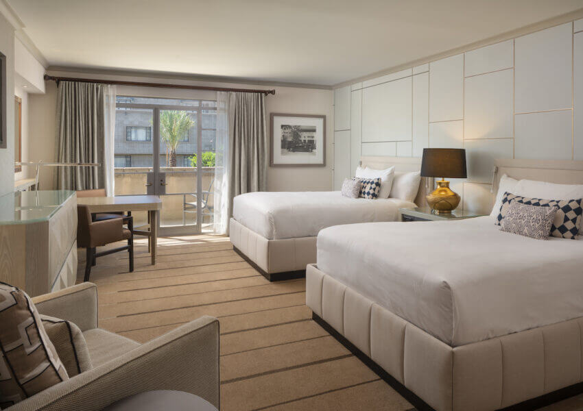 A modern hotel room with two large white beds, a seating area with a chair and desk, a lamp on a bedside table, and floor-to-ceiling windows covered by curtains. The room is well-lit by natural light, with light-colored walls and carpet.