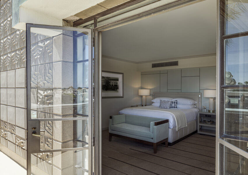 View into a modern bedroom through open French doors. The room features a large bed with white linens, a light blue bench at the foot, bedside tables with lamps, and a framed black and white photo on the wall. The exterior has a textured concrete facade with glass accents.