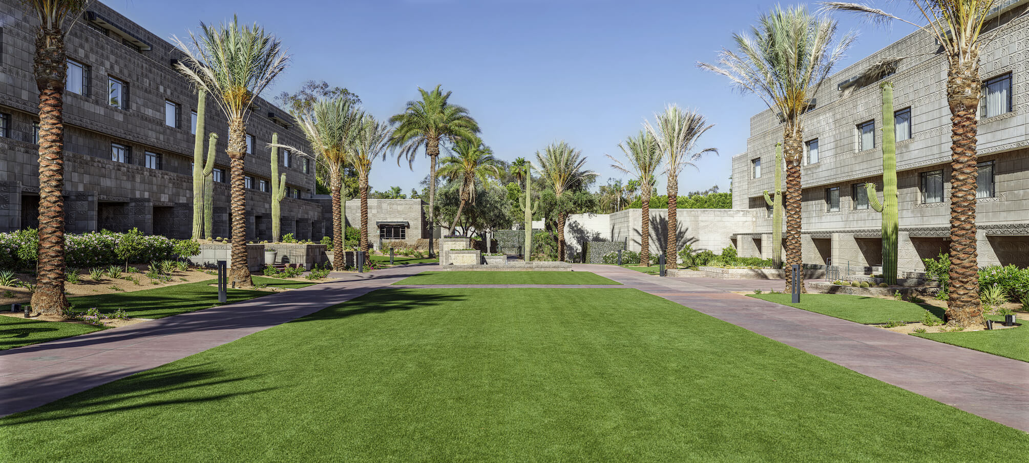 A landscaped courtyard with a wide expanse of green lawn is bordered by sidewalks and palm trees. Surrounding the courtyard are modern buildings with stone facades. In the background, there are more tall palm trees and shrubs under a clear blue sky.