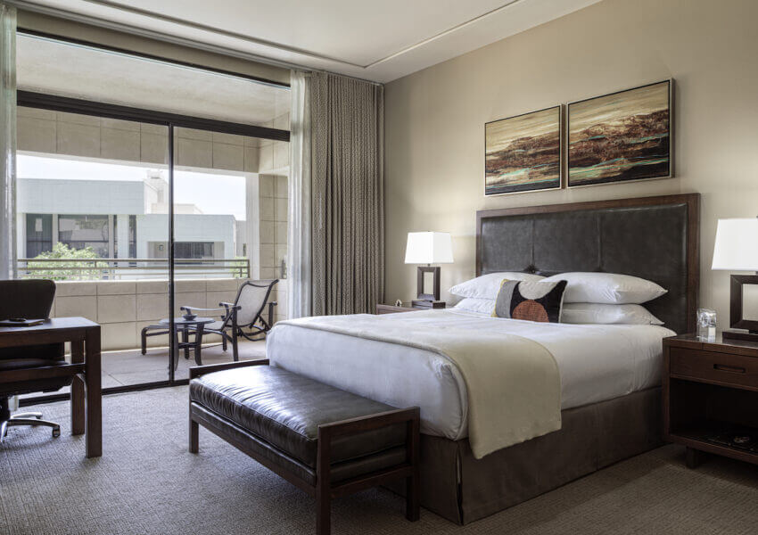 A modern hotel room featuring a king-sized bed with a padded headboard and bench at the foot. Two bedside tables with lamps, a desk with a chair, and a balcony with patio furniture are also visible. The walls are decorated with two landscape paintings.