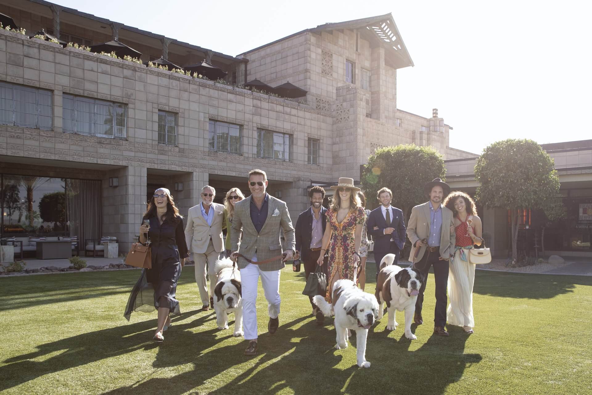 A group of nine people, some holding glasses, walk on a green lawn with two large fluffy dogs. They are dressed in stylish attire, standing in front of a large, elegant building with a modern architectural design. The pet-friendly scene is bright and sunny.