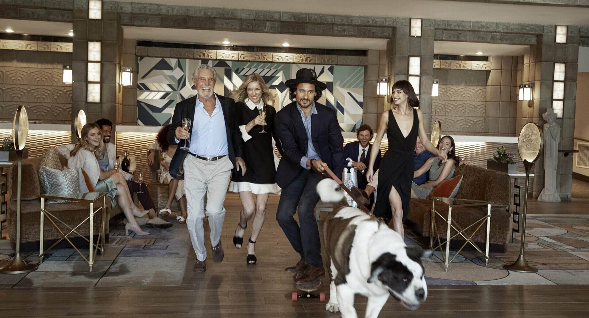 A joyful group of people in stylish attire follows a large Saint Bernard dog on a skateboard in an elegant room with modern decor. They seem to be celebrating their resort membership, with some holding glasses of wine, smiling, and appearing to be in high spirits.