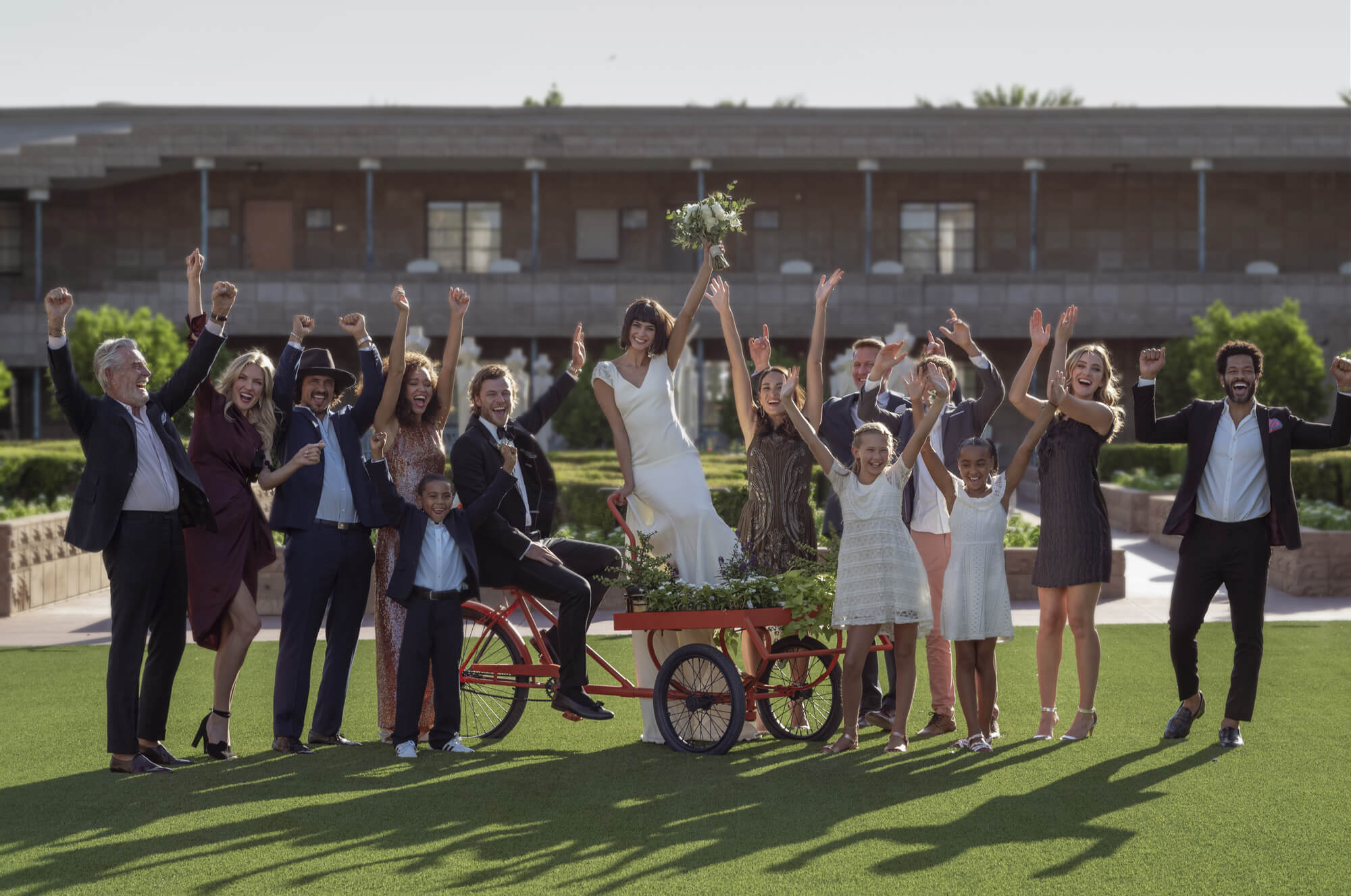 A joyful group of people stands on a grassy area in front of a building, raising their arms in celebration. Amid the wedding festivity, a woman in a white dress stands on a cart and holds a bouquet aloft. The group includes adults and children, all appearing happy and exuberant.