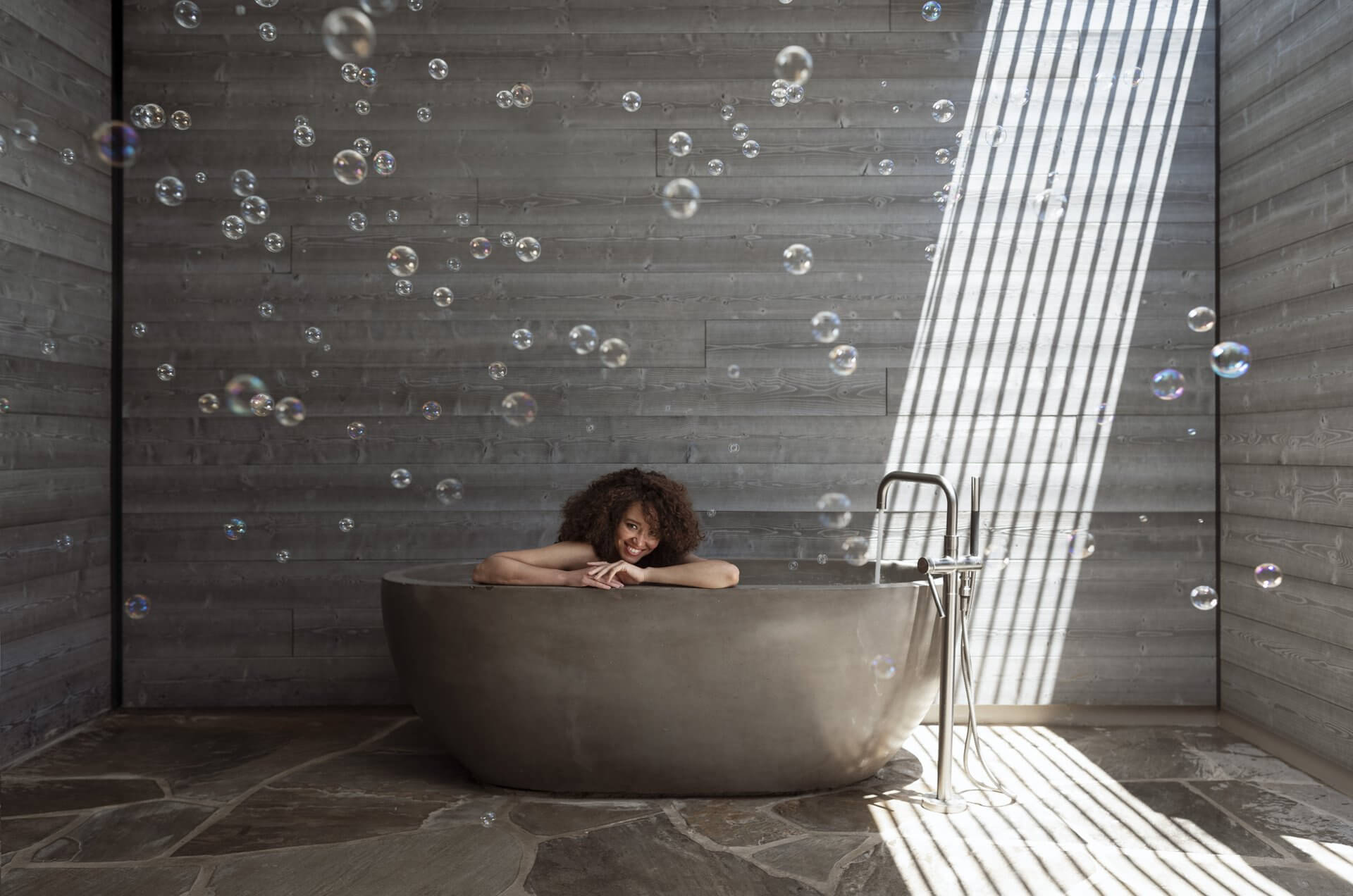 A person with curly hair relaxes in a large, modern stone bathtub in a room with wooden paneled walls and stone flooring. Sunlight streams in through a window with blinds, casting striped shadows, while bubbles float around the room, creating a perfect spa & wellness experience.