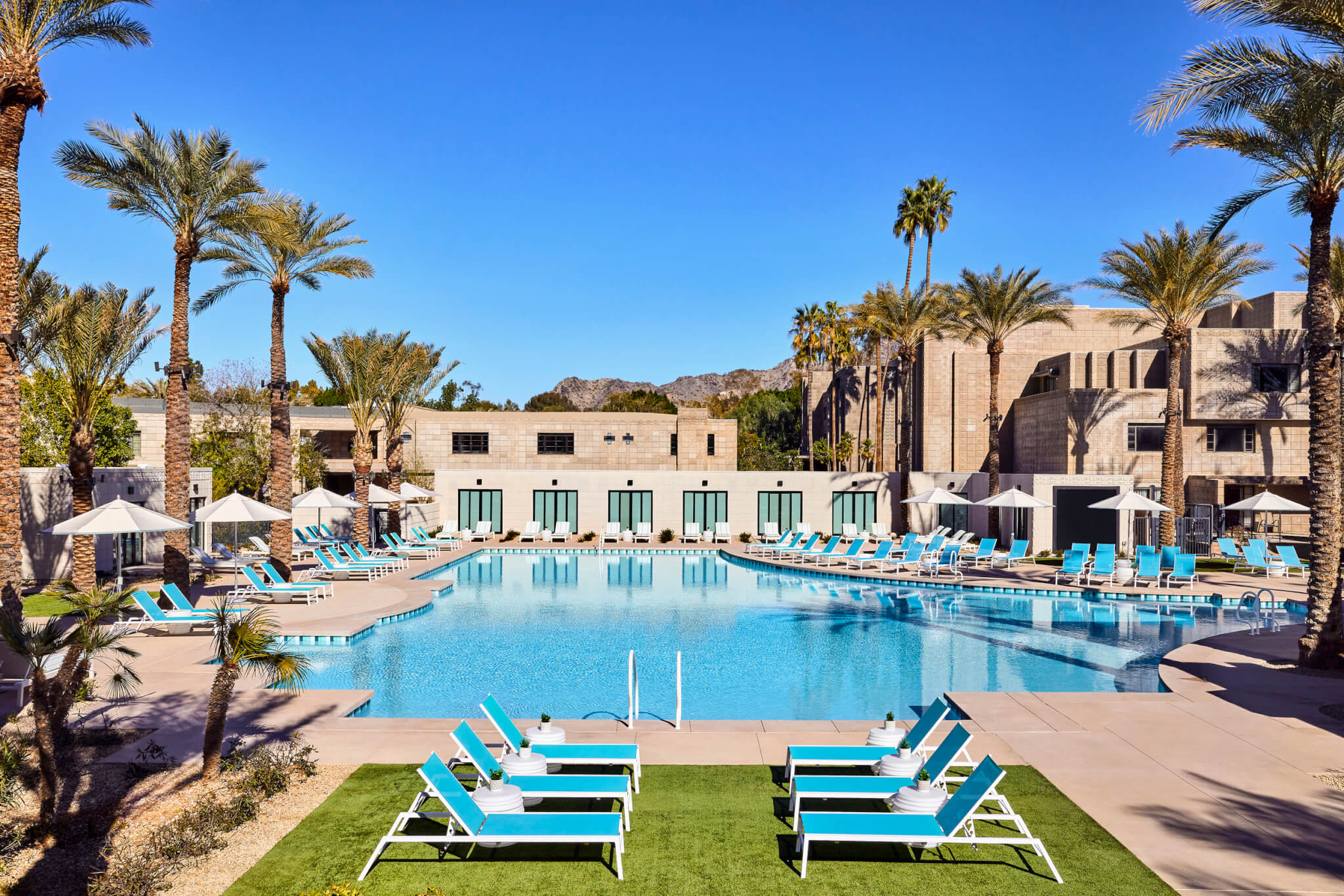 A luxurious paradise pool surrounded by lounge chairs with turquoise cushions, shaded by tall palm trees. The pool area is flanked by modern building architecture, and mountains are visible in the background under a clear blue sky.