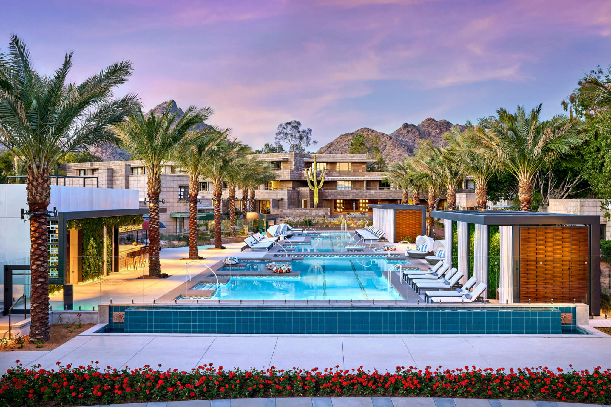 A luxurious outdoor pool area at the modern Arizona Biltmore resort under a purple and pink sky at sunset. The pool is surrounded by lounge chairs, cabanas, palm trees, and neatly landscaped areas with red flowers. Mountains can be seen in the background.