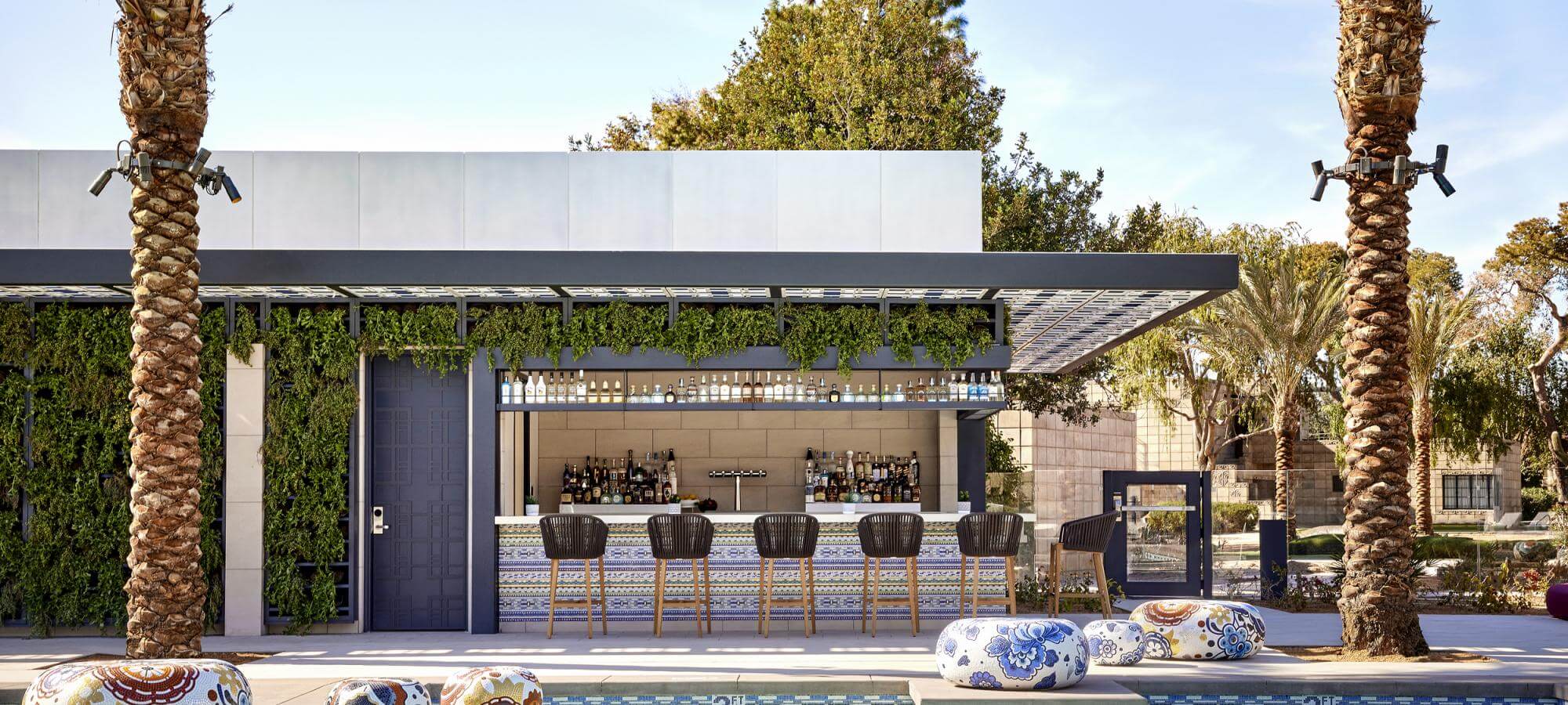 Outdoor bar with a canopy and green vines hanging from the roof edge. It has a row of high-back bar stools, a counter, and shelves holding bottles like treasures of history. Surrounding the bar are palm trees, colorful cushioned stools, and a small pool in the foreground. Sunny day.