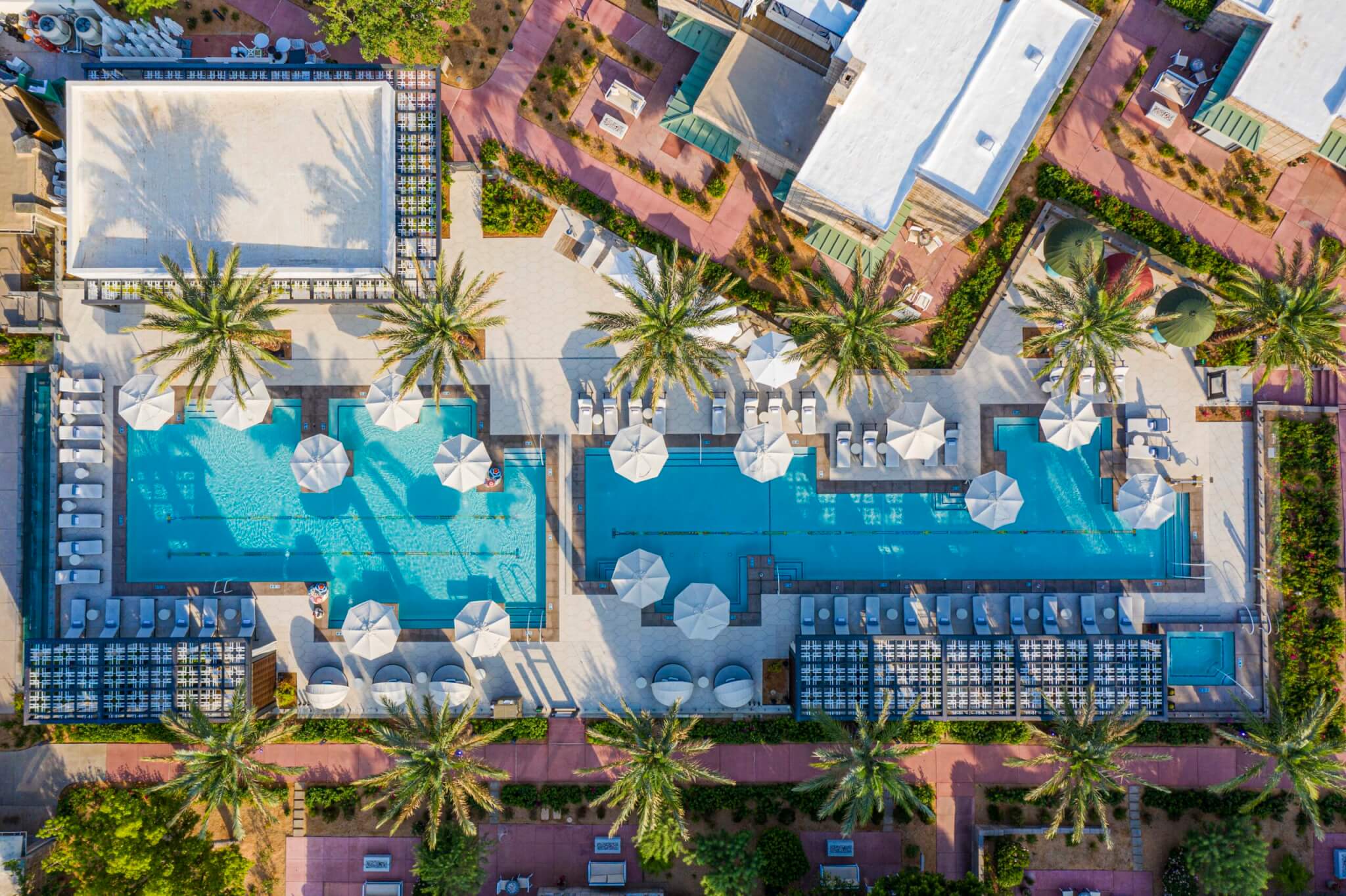 Aerial view of the luxurious Arizona Biltmore Pools at a resort. The scene features two large rectangular pools surrounded by palm trees, lounge chairs, and umbrellas. Adjacent buildings with white roofs and landscaped walkways are visible in the background.