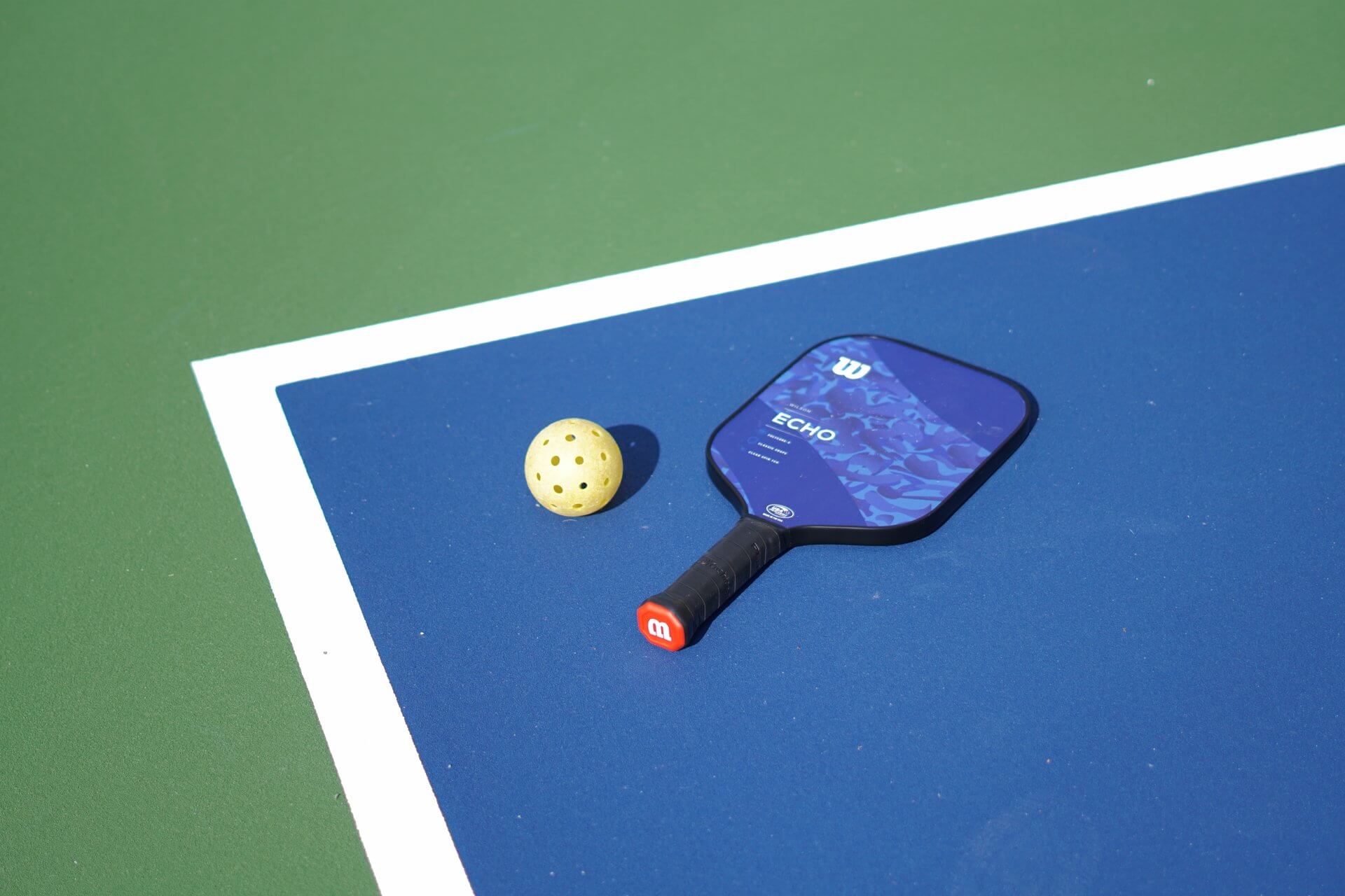 A blue pickleball paddle and a yellow perforated pickleball lie on a blue and green hard court next to a white boundary line, indicating the game is played outdoors under bright sunlight—perfect conditions for our upcoming pickleball clinic.