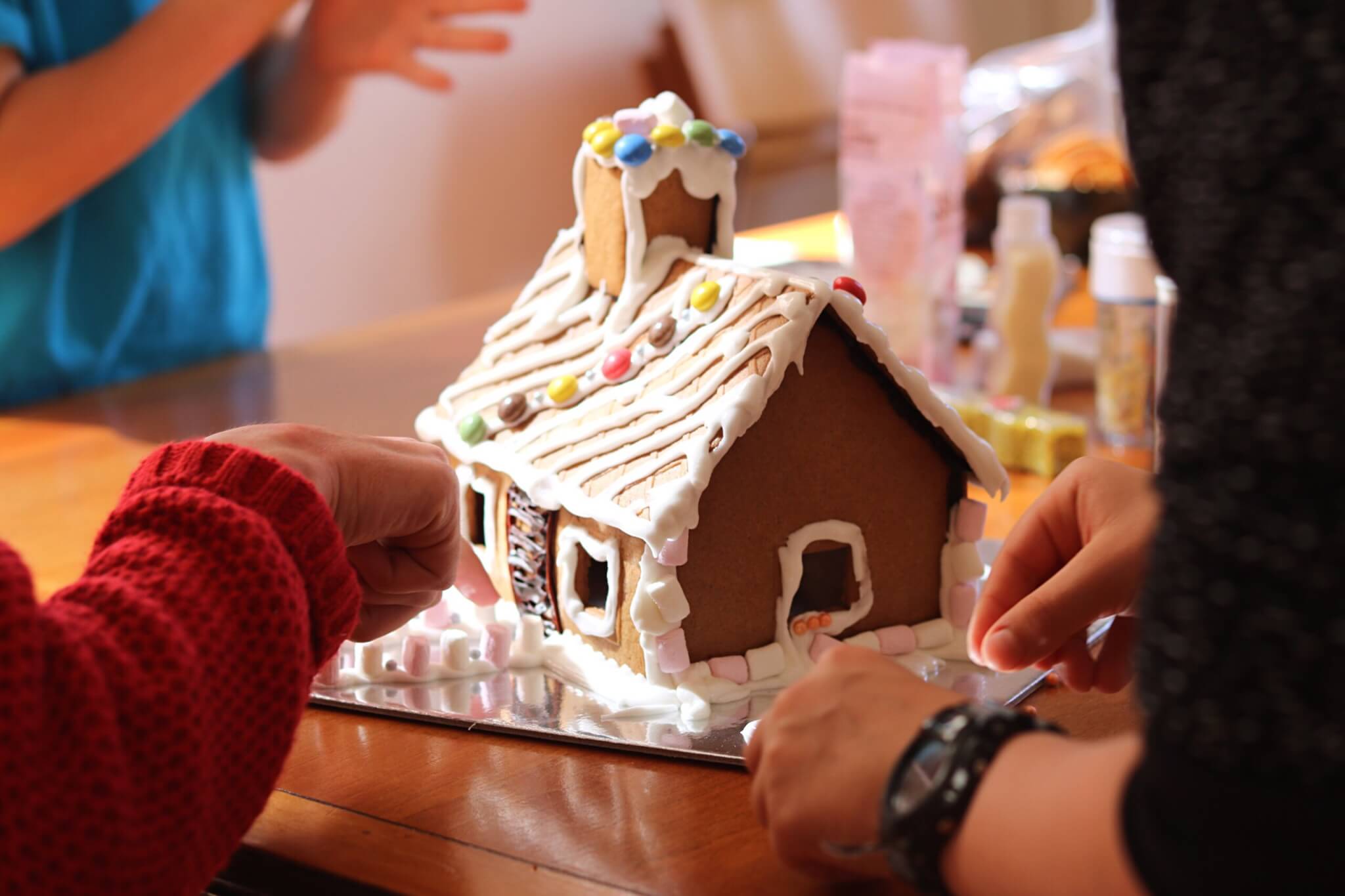 Three people decorate a gingerbread house with white icing and colorful candy on a table, perhaps as a sweet addition to their Thanksgiving Dinner To Go. Their hands place candies on the roof, while marshmallows line the base of the house.