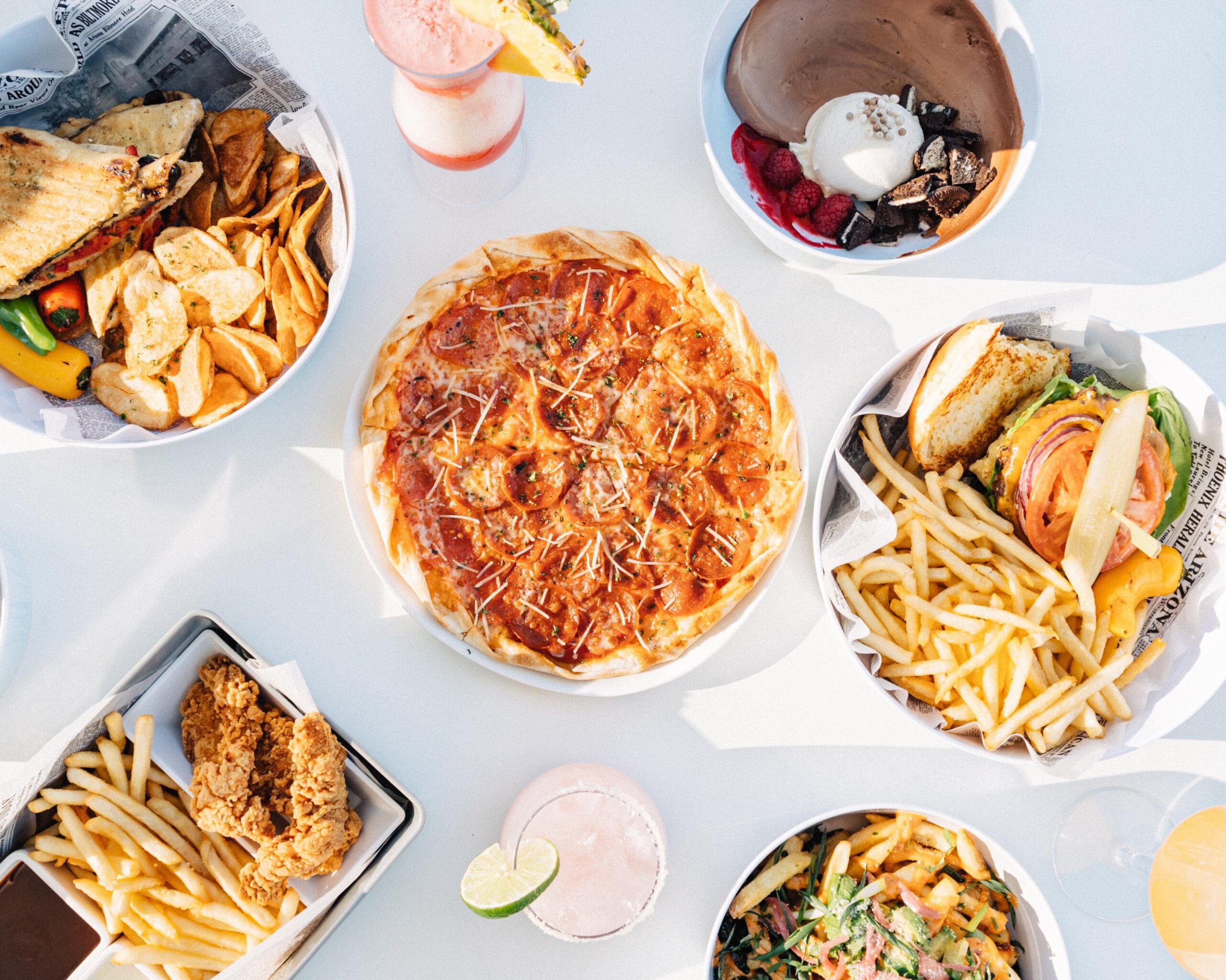 A table with various foods including pepperoni pizza, French fries, fried chicken, a sandwich, salad, a chocolate dessert bowl, and colorful drinks on a white surface.