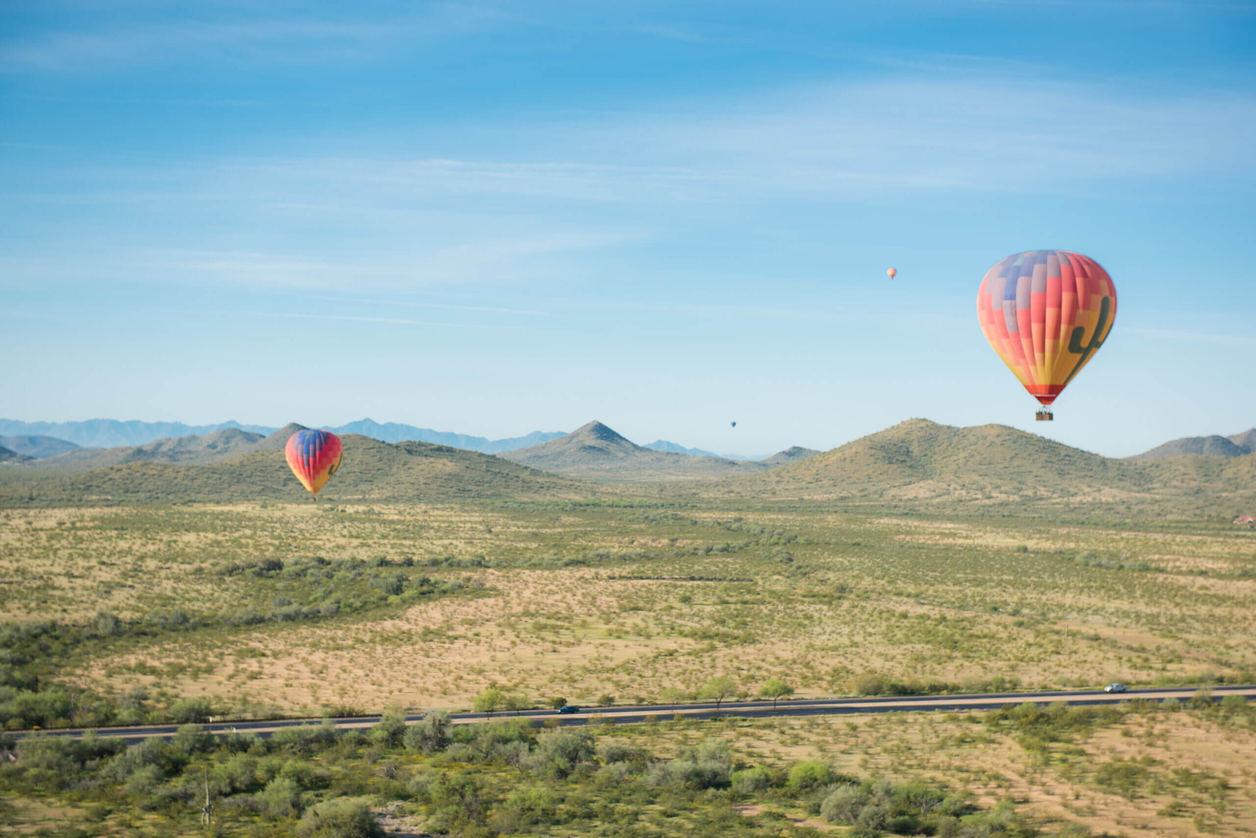 A scenic landscape with two colorful hot air balloons floating above a vast, arid terrain invites you to explore. The terrain is dotted with sparse vegetation and crossed by a road with a few vehicles. Rolling hills and distant mountains are visible under the clear blue sky.