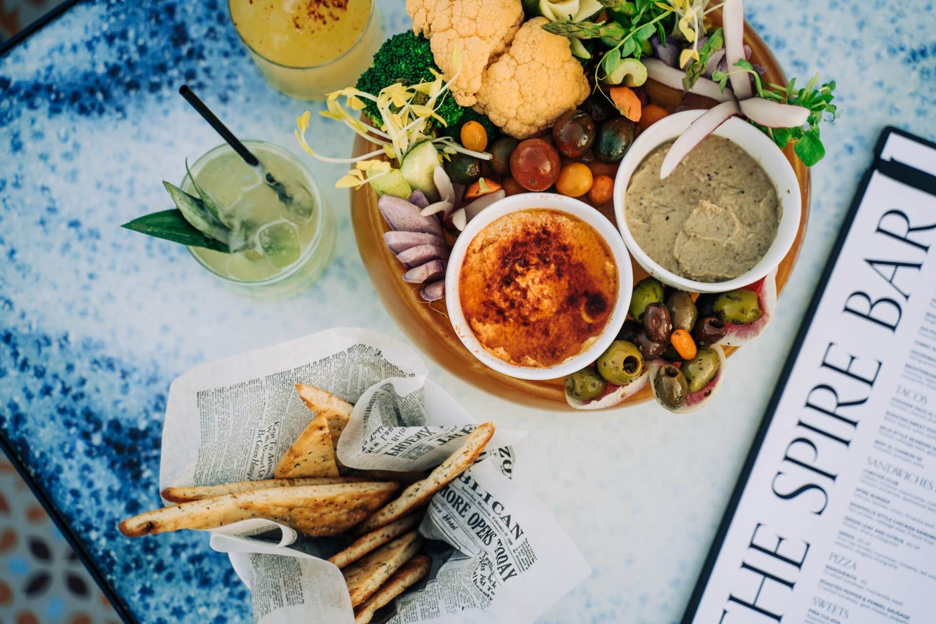 A top view of a vibrant Mediterranean platter featuring various dips, olives, cauliflower, and other vegetables served on a wooden board. On the side, a basket holds pita bread, and drinks are placed nearby on a speckled tabletop with a menu labeled "The Spire Bar.