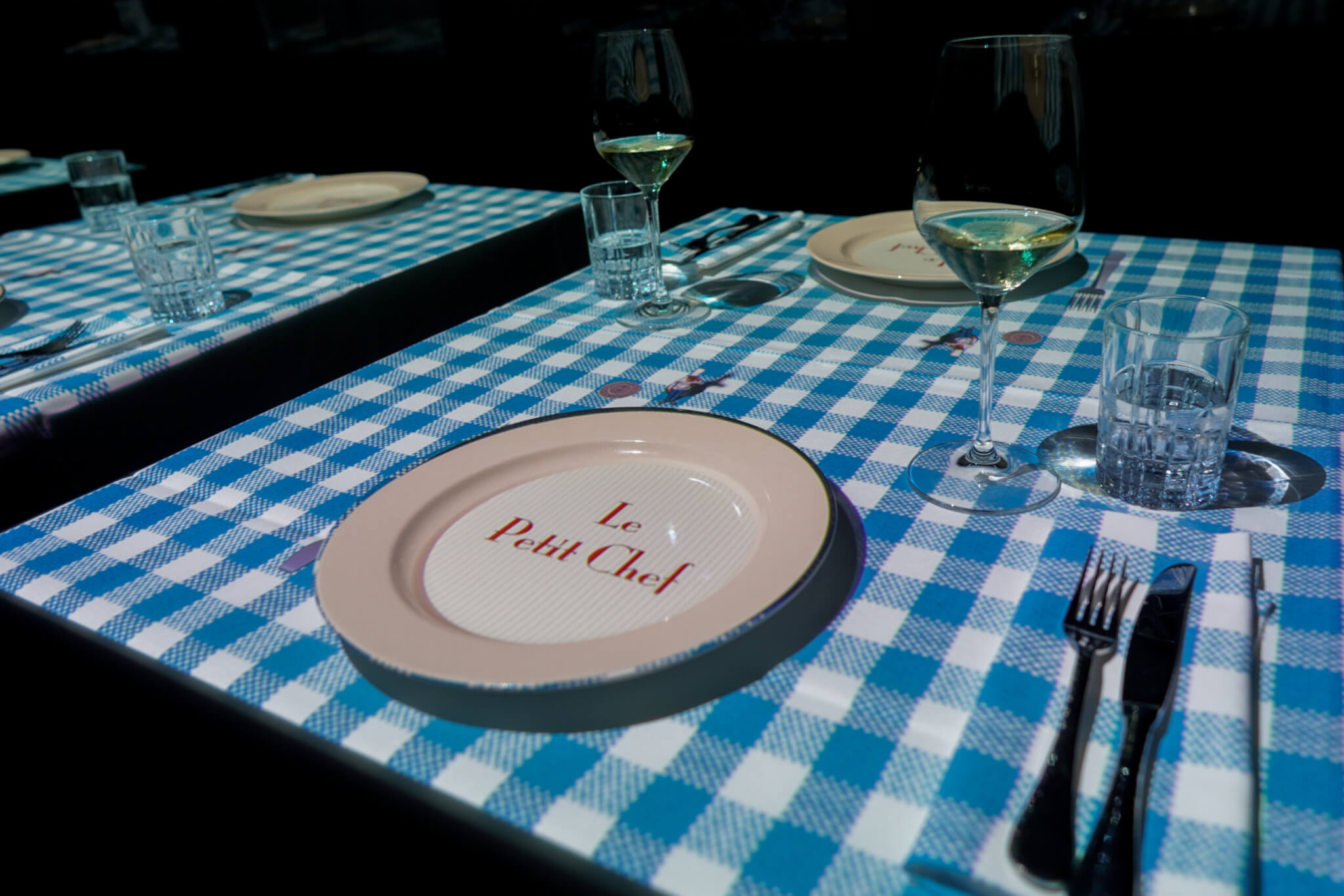 A table set for two with blue and white checkered tablecloths, plates labeled "Le Petit Chef," wine glasses filled with white wine capture the Last Tastes of Summer, water glasses and cutlery neatly arranged.