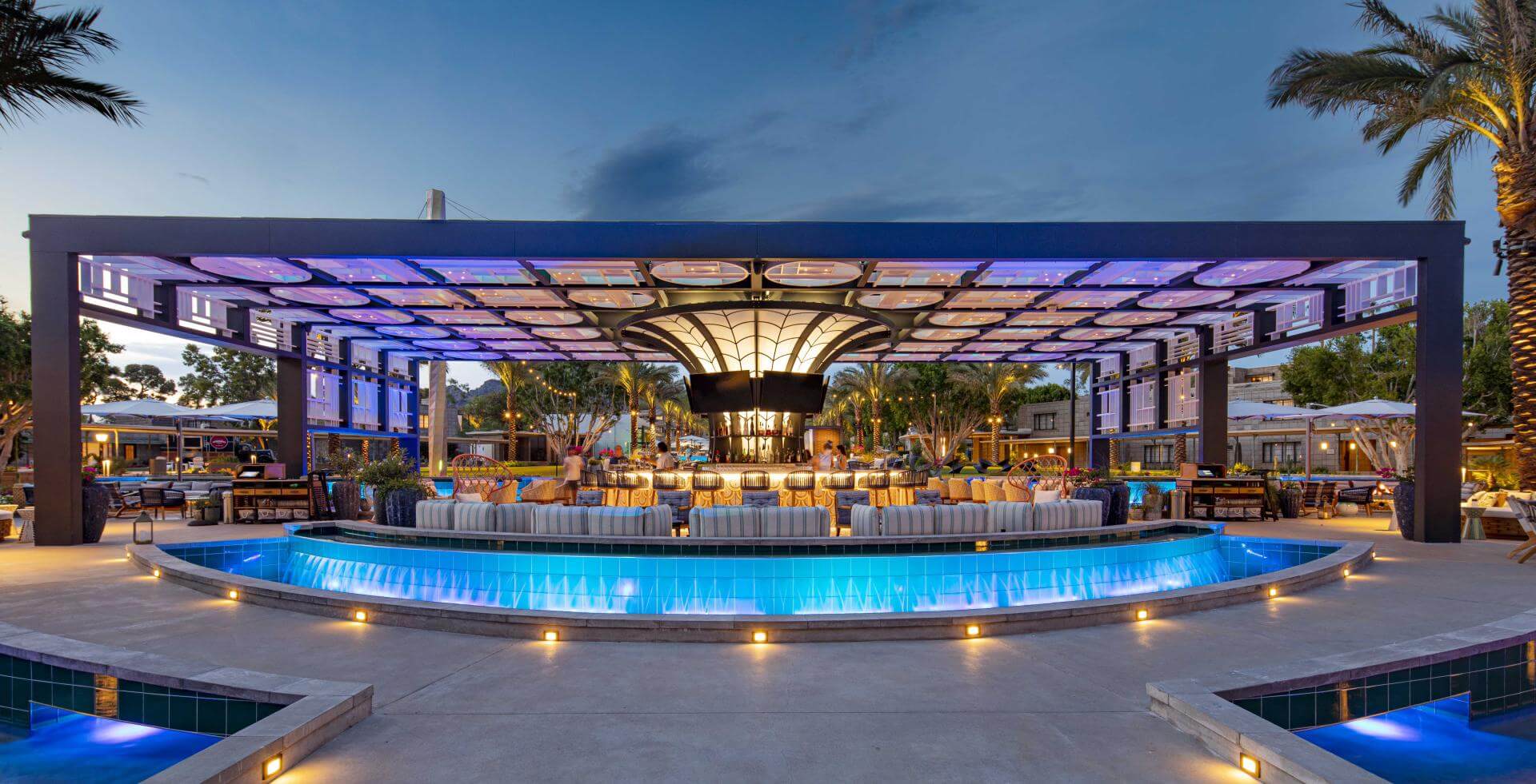An outdoor spire bar and lounge area under a modern, open-frame structure, illuminated with blue and white lights in the evening. The area is surrounded by a curved pool and furnished with tables and chairs, set amidst palm trees and a serene atmosphere.