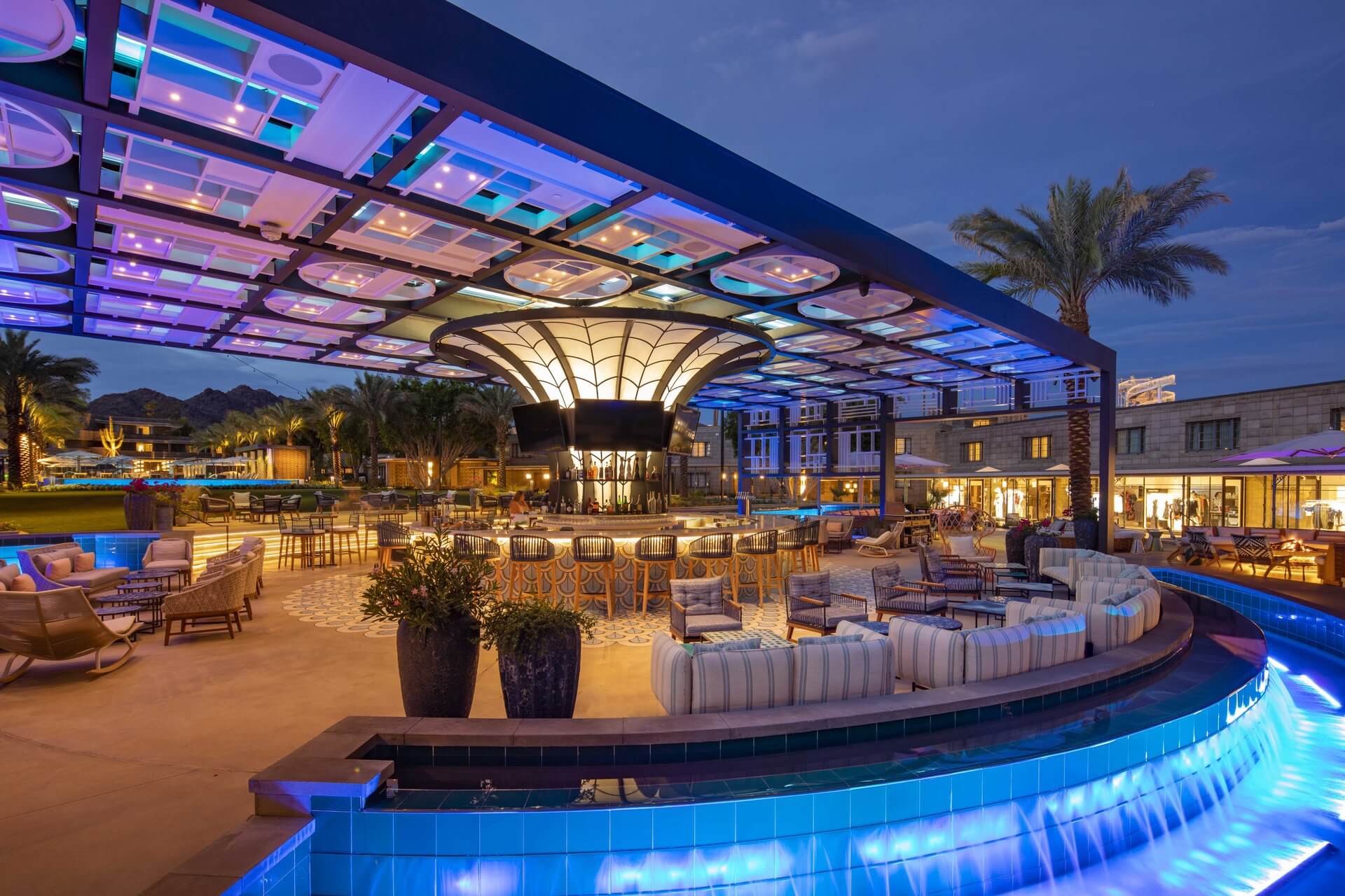 An outdoor lounge area at night, featuring a central bar with stools under a modern, illuminated canopy. Surrounding seating includes wicker furniture and cushioned chairs. Palm trees, upscale buildings, and a curved pool with a blue tile design complete the scene.