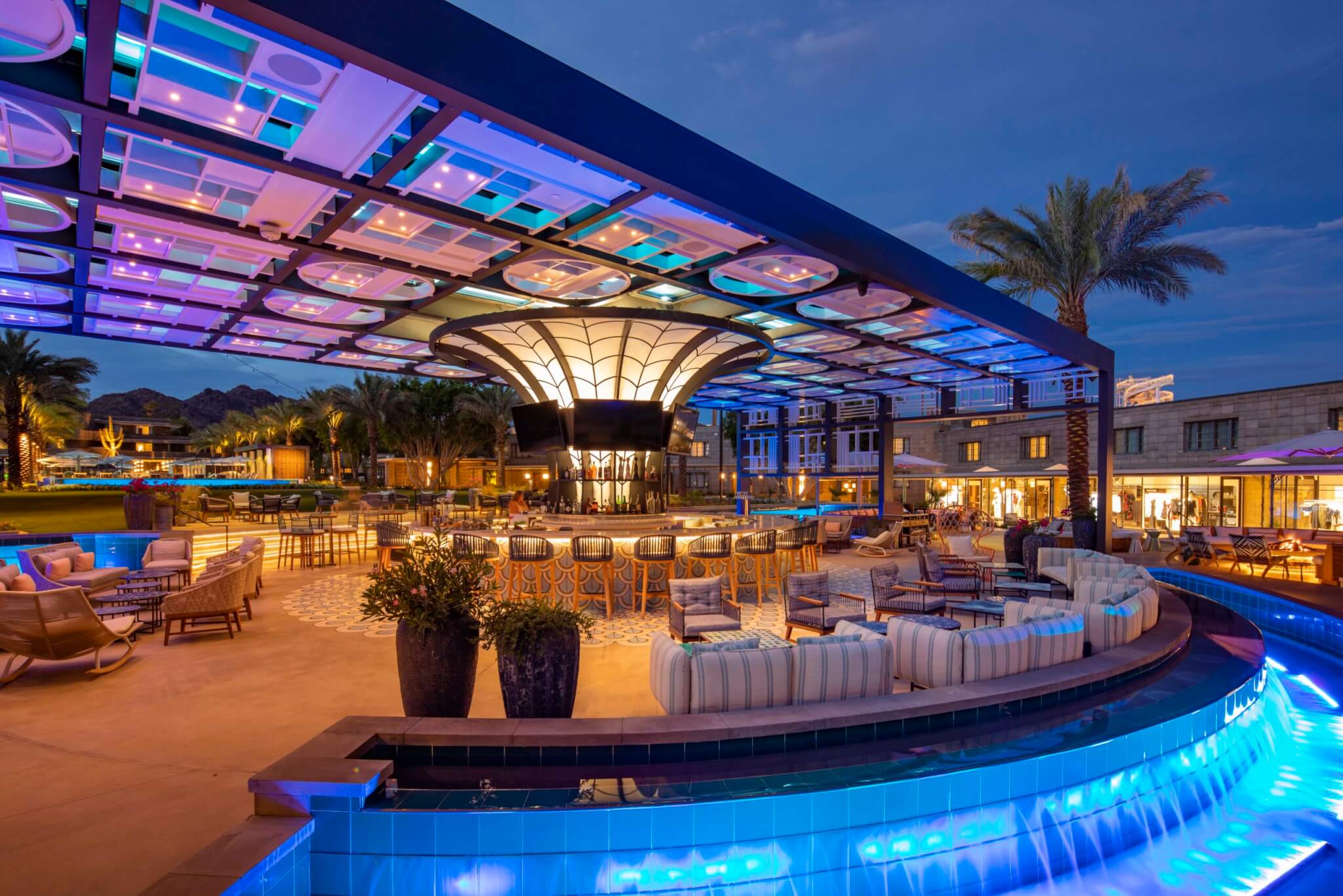 Outdoor bar and lounge area lit with colorful, modern lighting under a geometric canopy, surrounded by a pool with blue lights, palm trees, and elegant seating at a resort in the evening.