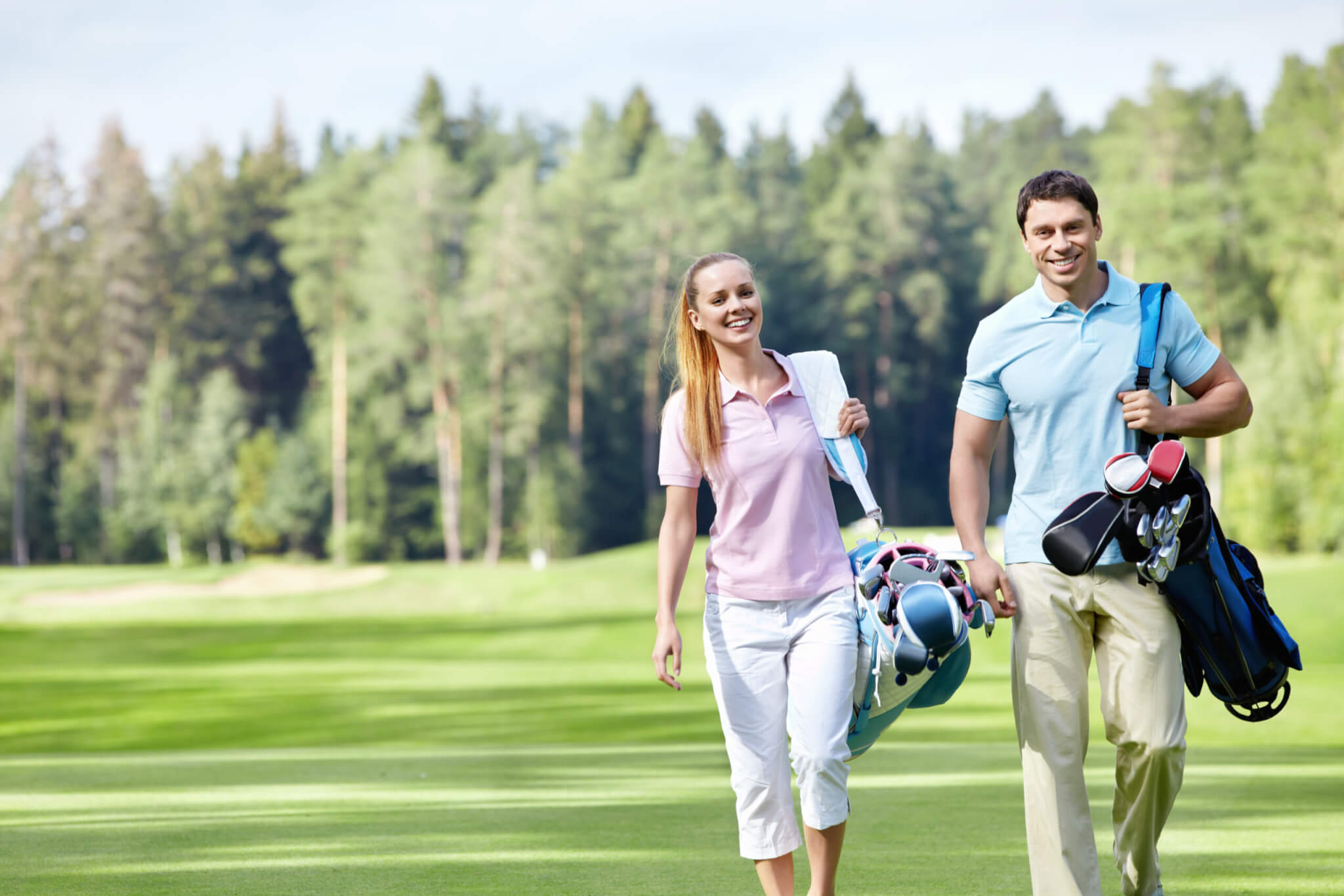 A man and a woman walk on a golf course, smiling and carrying golf bags. They are dressed in casual golf attire, with the woman in a pink polo and white pants, and the man in a light blue polo and beige pants. Trees and green grass of the pristine golf course are visible in the background.