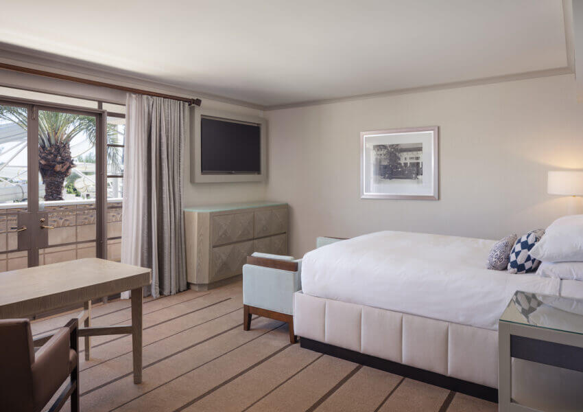 A modern hotel room featuring a large bed with white linens, a wooden desk and chair, a flat-screen TV on the wall, and a framed picture above a dresser. A large sliding glass door with curtains opens to a balcony with a view of palm trees.