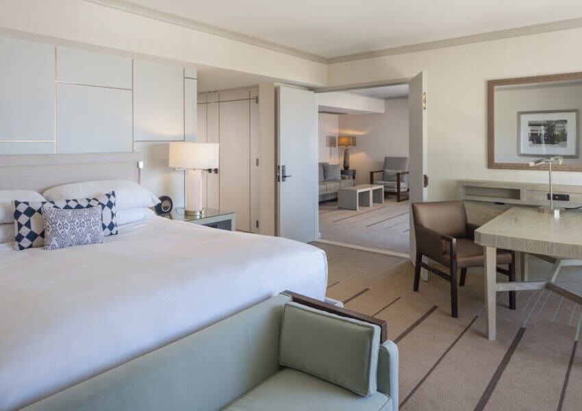 A spacious hotel room with a large bed adorned with white linens and decorative pillows. There is a small seating area with a green sofa, a wooden desk and chair, and a lamp. In the background, a separate living area is visible with additional seating.