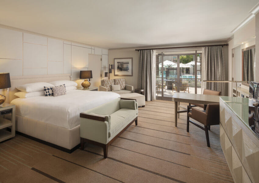 A spacious hotel room with a large king-sized bed, flanked by two bedside tables with lamps. A light green bench is at the foot of the bed. The paradise poolside room features a desk, chair, an armchair with an ottoman, and a TV. Large windows reveal a balcony overlooking the pool area.