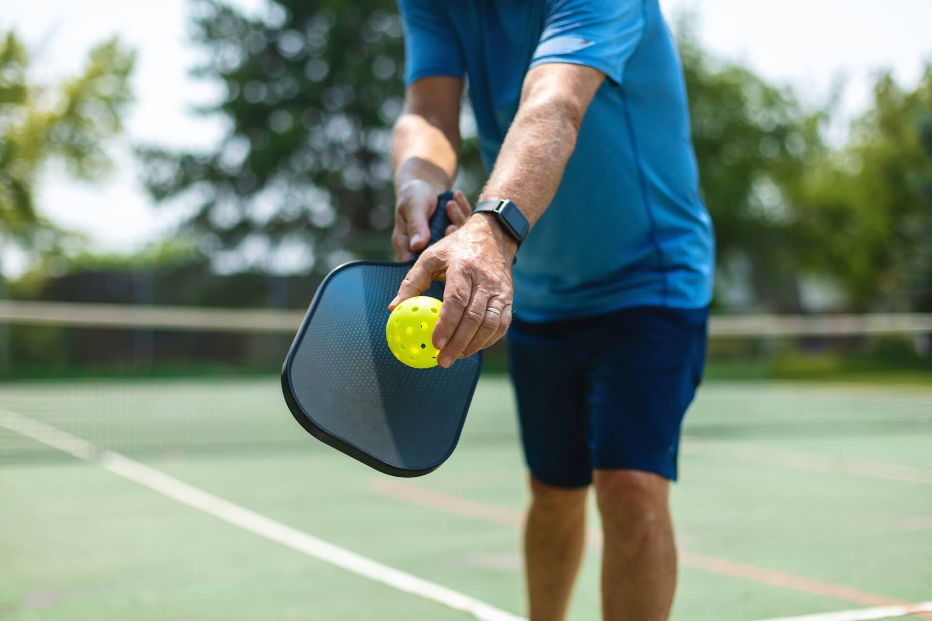 A person in a blue shirt and shorts is holding a yellow perforated ball with one hand and a pickleball paddle in the other, positioned to serve on an outdoor court—an ideal activity for those enjoying their resort membership. The background includes a blurred view of the net and surrounding greenery.