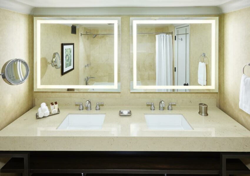 A modern bathroom with a double-sink vanity featuring two backlit mirrors. The countertop holds toiletries, towels, a soap dish, and a metal tissue holder. A bathtub with a white shower curtain and a framed picture are visible in the background.