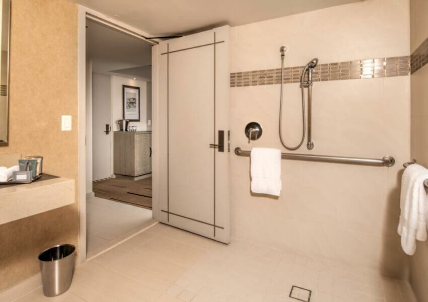 A hotel bathroom with beige walls and tile flooring designed for accessibility. It features an accessible shower with a roll-in entry, handheld showerhead, and grab bars, while a white towel hangs from one of the bars. The vanity holds towels and toiletries, and there is a waste bin on the floor. An open door reveals the adjacent room.