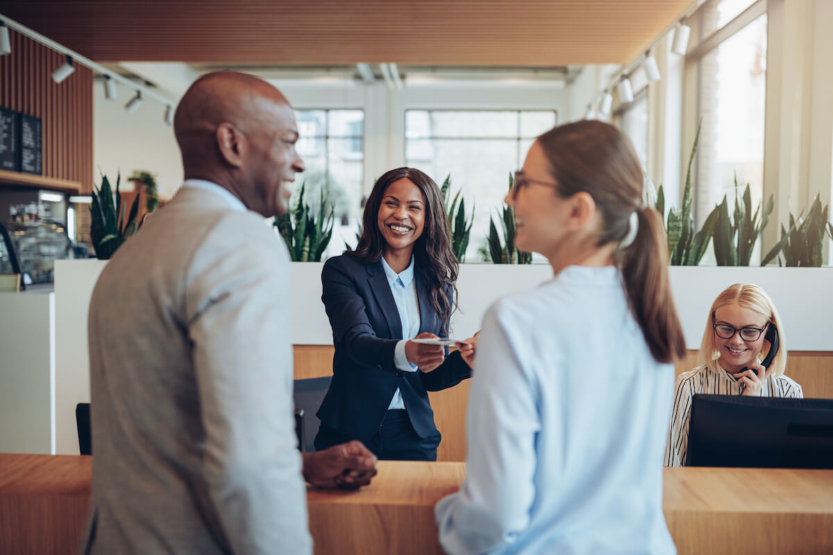 A smiling businesswoman standing behind a reception desk hands a card to a client, discussing potential careers. Another woman with glasses and a ponytail talks to the client, while a receptionist in the background works on a computer. The setting appears to be an office.
