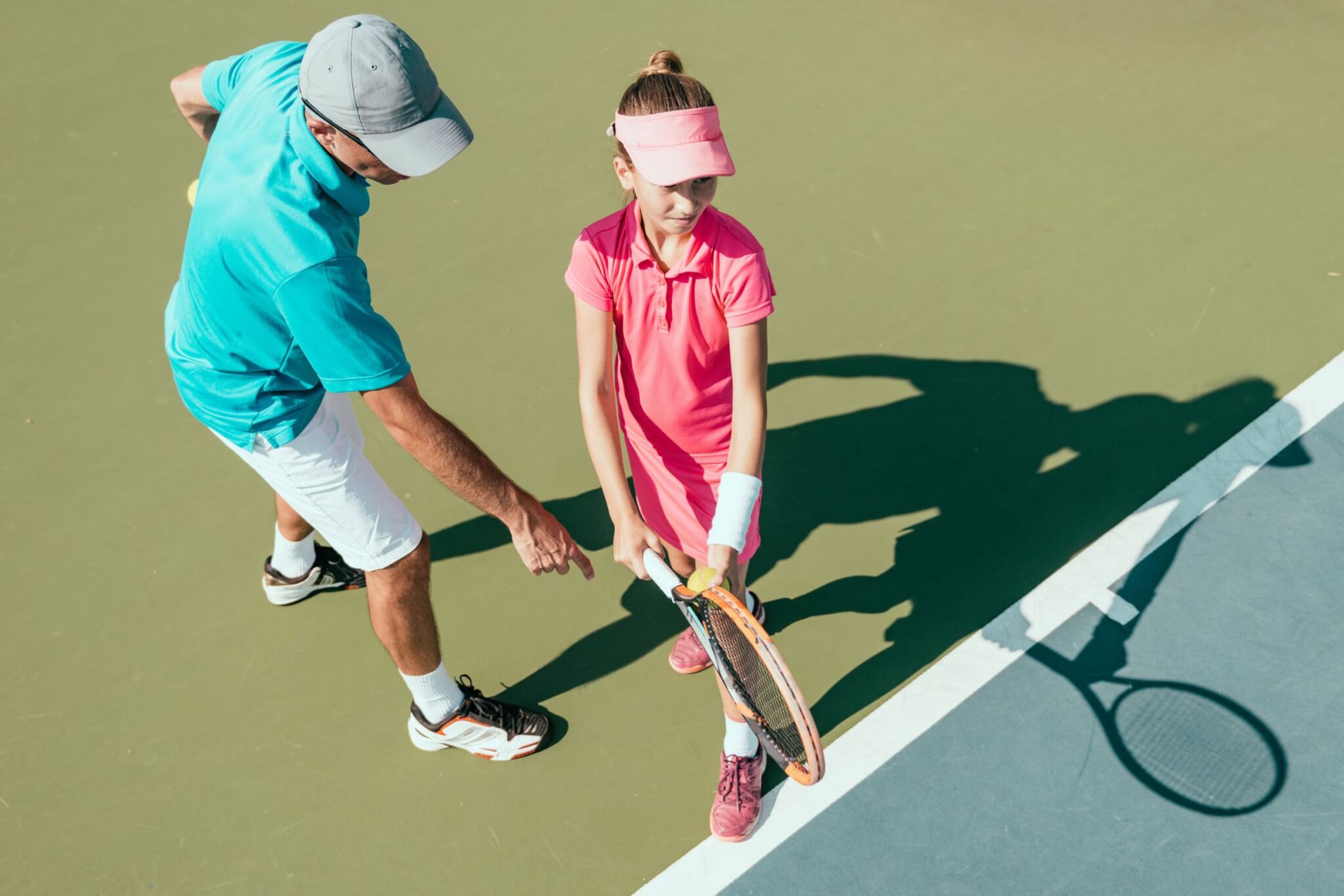 A tennis coach in a blue shirt and white shorts instructs a young girl in a pink outfit and visor on a tennis court. The coach points towards the girl's racket as she holds it, preparing to hit a ball.