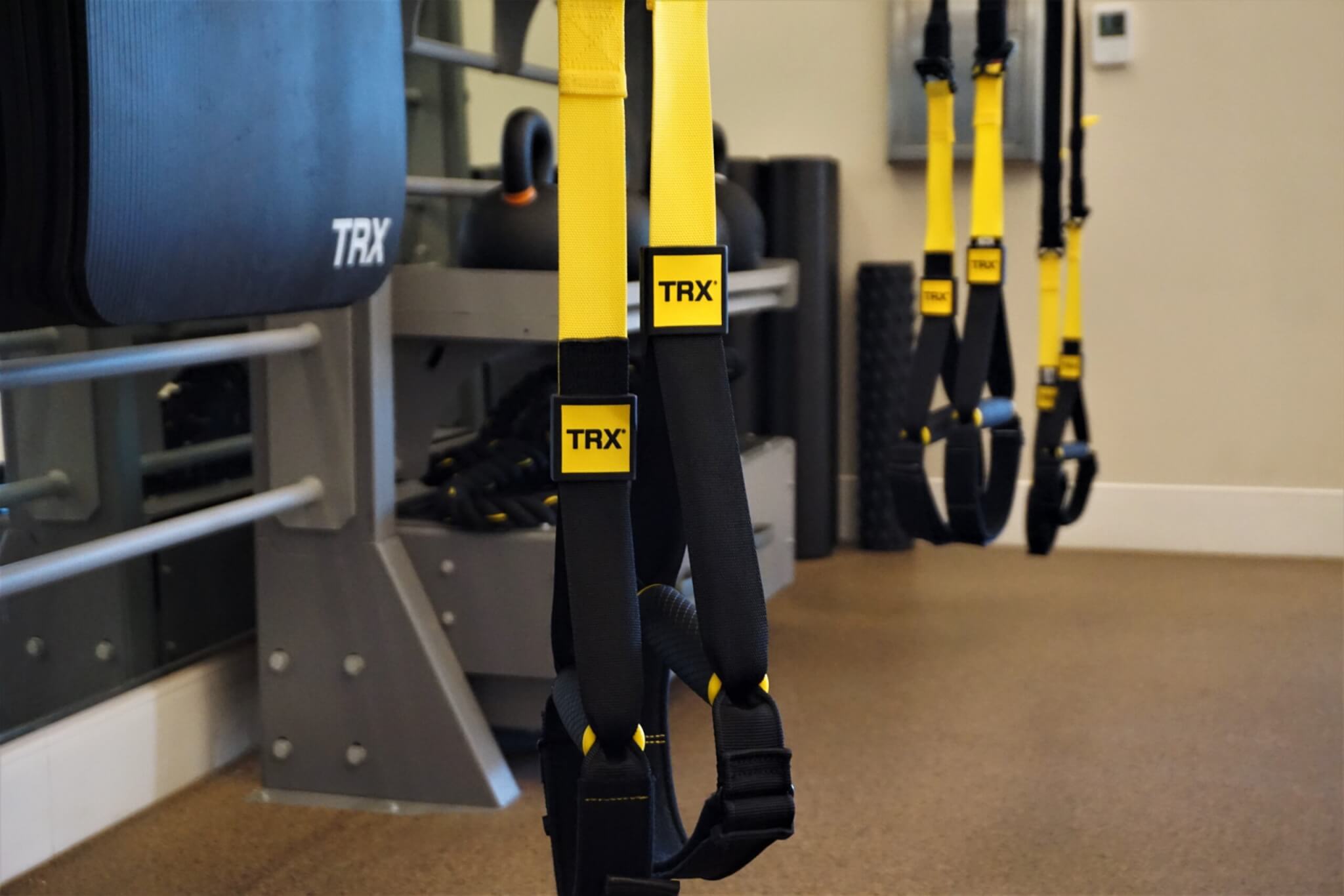 Close-up of yellow and black TRX suspension straps hanging in a gym, with exercise equipment and a mirror in the background.