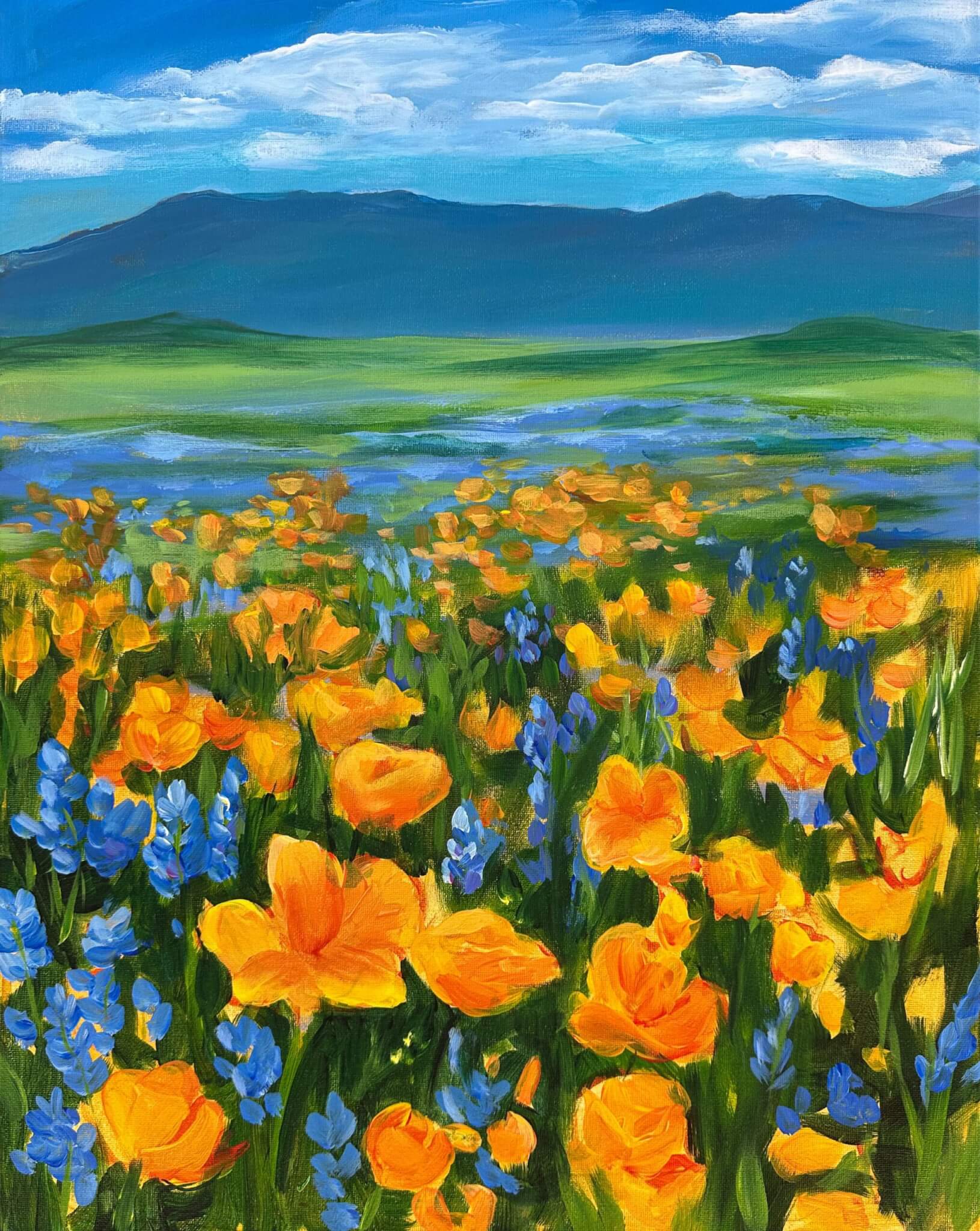 A vibrant field of orange and blue wildflowers stretches toward distant green hills and dark mountains under a bright blue sky with fluffy clouds.