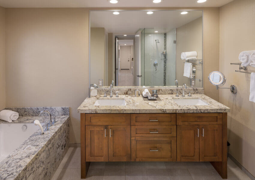 A spacious modern bathroom with granite countertops featuring a double sink vanity, a large mirror, and overhead lighting. To the left is a built-in bathtub with a granite surround. A glass-enclosed shower is visible in the background. Towels are neatly hung.