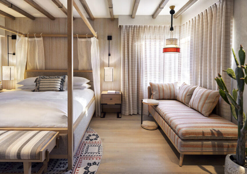 A cozy cottage suite bedroom featuring a wooden four-poster bed adorned with white linens, a patterned bench at the foot, and a nightstand with a lamp. A striped chaise lounge sits near a window with sheer curtains, complemented by indoor plants and a woven rug.