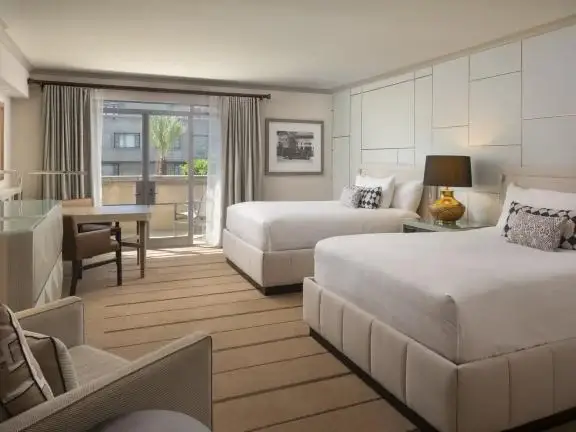 A luxurious hotel room features two large beds with white linens, a small table and chair near a window, and modern decor. A glass door opens to a balcony with a view of the city. Neutral tones accent the room, enhanced with warm lighting from a bedside lamp.
