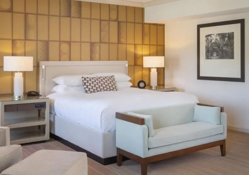 A modern hotel room invites you to stay with a large, neatly-made bed adorned with a decorative pillow. Flanking the bed are two side tables with lamps. A light-colored armchair and ottoman sit in the foreground, while a cushioned bench rests at the foot of the bed. A large mirror and artwork adorn the walls.